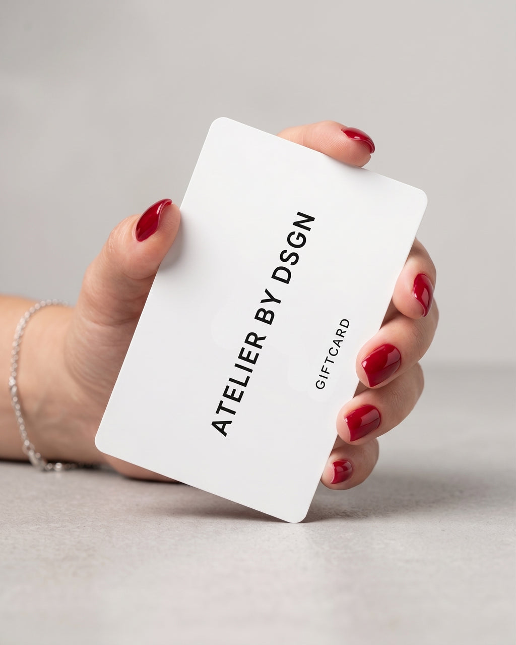 A hand with red nail polish holds a white Atelier By DSGN gift card against a neutral light gray background.