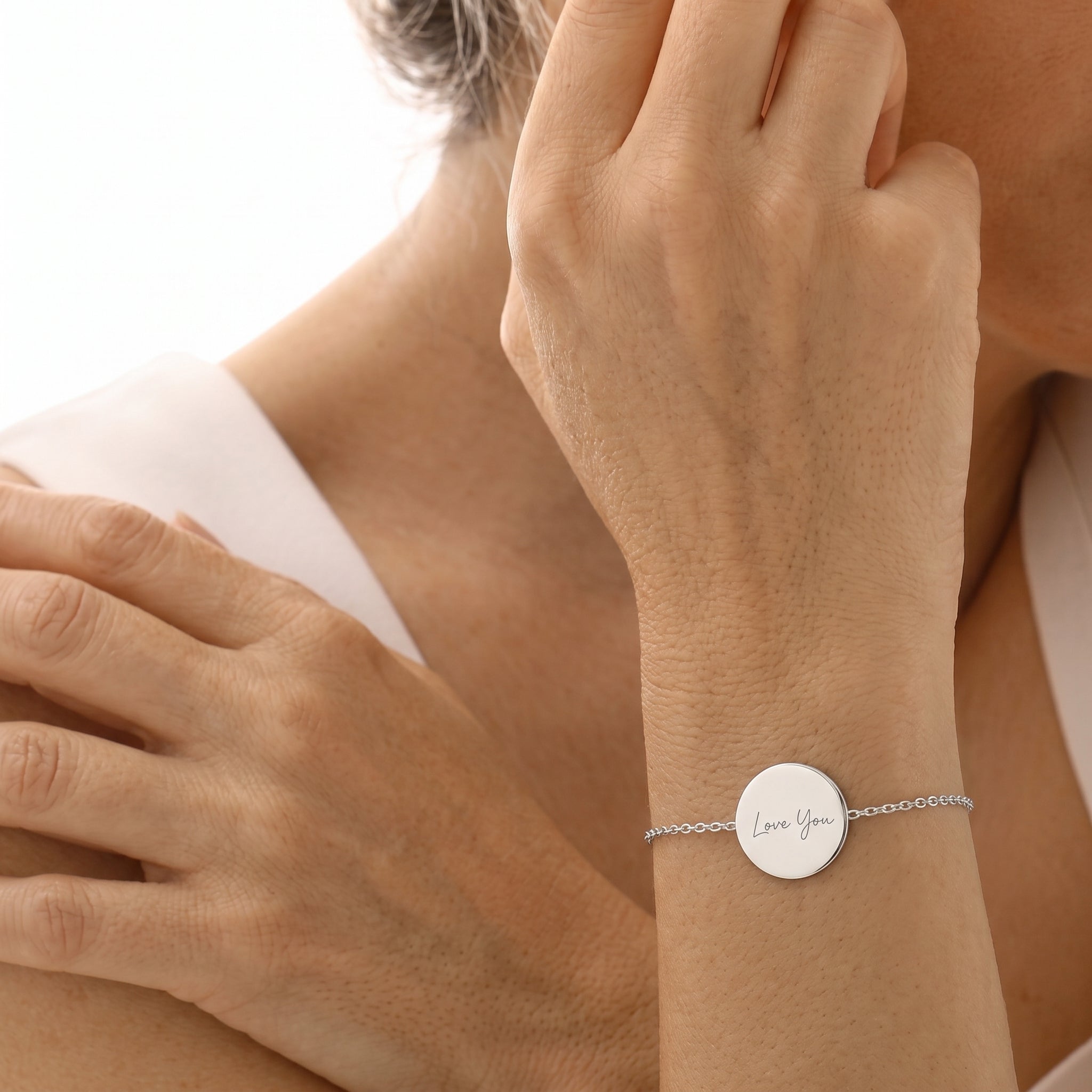 A close-up of a delicate silver chain bracelet with a round silver disc engraved with the words 'Love You', worn on a woman's wrist.  Meaningful custom jewelry from Atelier By DSGN