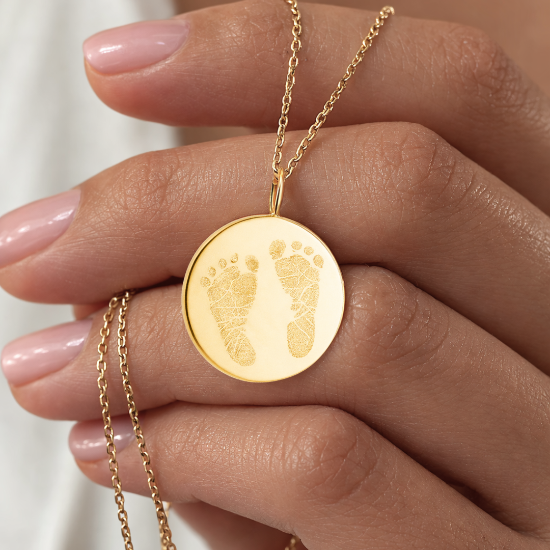 Close-up of custom engraved 18k gold vermeil 16mm coin pendant necklace resting in female palm with nude nails, showcasing intricate personalised double newborn baby footprints with detailed toe and sole texture precision diamond-head engraved on warm lustrous gold disc on delicate gold diamond-cut cable chain – Atelier By Dsgn luxury custom footprint coin necklace cherished gift for new mum, mothers day, baby shower, birth announcement or treasured newborn family heirloom necklace