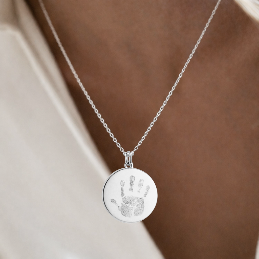 Custom engraved 925 sterling silver baby handprint coin pendant necklace worn at neckline on female model in cream top, personalised baby handprint with intricate finger and palm detail precision diamond-head engraved on polished round disc on delicate diamond-cut cable chain – Atelier By Dsgn custom handprint pendant jewellery heartfelt gift for new mum, mothers day, baby shower, new baby arrival or treasured newborn family keepsake necklace