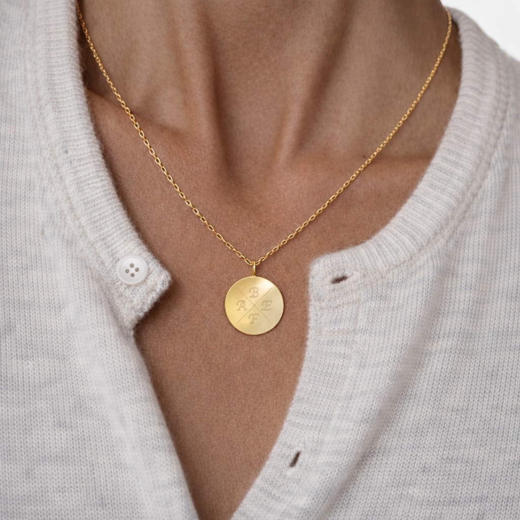 Custom engraved 18k gold vermeil 16mm coin pendant necklace worn at neckline on female model in cream cardigan, featuring personalised family initials "A, B, E, F" precision diamond-head engraved in elegant cross pattern on warm lustrous gold disc on delicate gold diamond-cut cable chain – Atelier By Dsgn luxury personalised initial coin necklace everyday wear gift for her, mothers day, family keepsake, anniversary or meaningful custom initials heirloom necklace