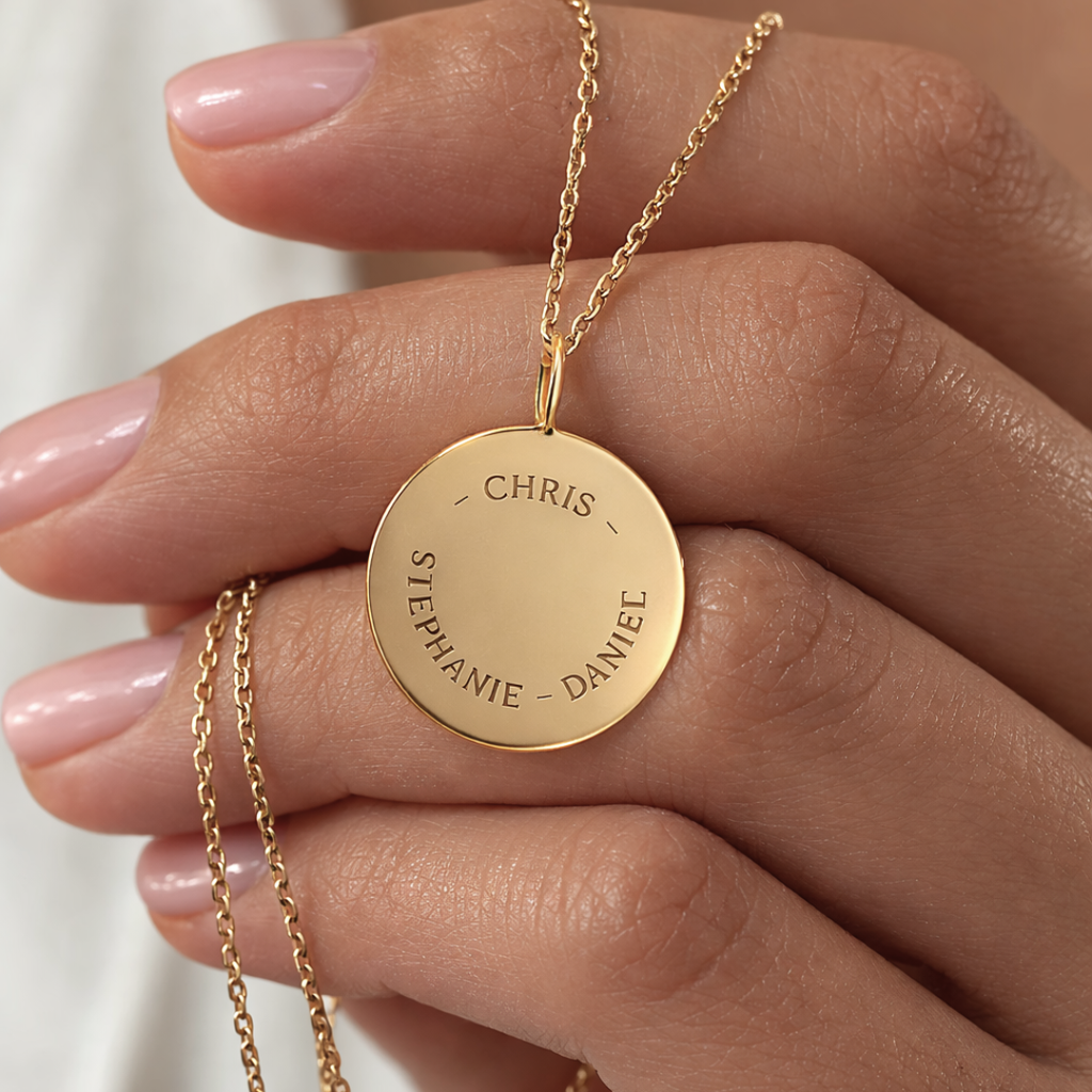 Close-up of custom engraved 18k gold vermeil 16mm coin pendant necklace held in female hand with nude nails, showcasing personalised family names "Chris, Stephanie, Daniel" precision diamond-head engraved in elegant curved layout on warm lustrous gold disc on delicate gold cable chain – Atelier By Dsgn luxury personalised text coin necklace meaningful gift for her, mothers day, family keepsake, anniversary or cherished custom names heirloom necklace