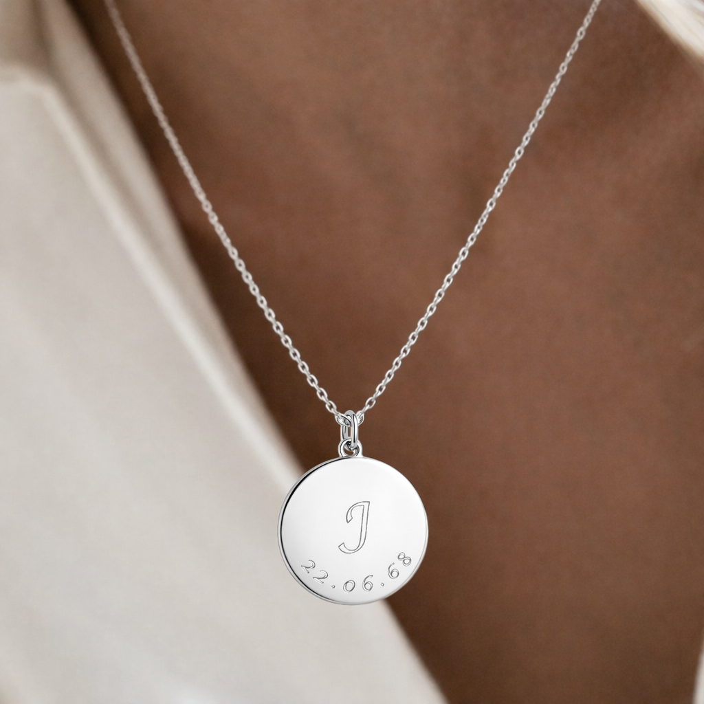Custom engraved 925 sterling silver 16mm coin pendant necklace worn at neckline on female model in cream top, personalised initial "J" with significant date "22.06.68" precision diamond-head engraved on polished round disc on delicate diamond-cut cable chain – Atelier By Dsgn personalised text pendant necklace elegant everyday wear gift for her, birthday milestone, mothers day, anniversary or cherished custom initial and date keepsake necklace