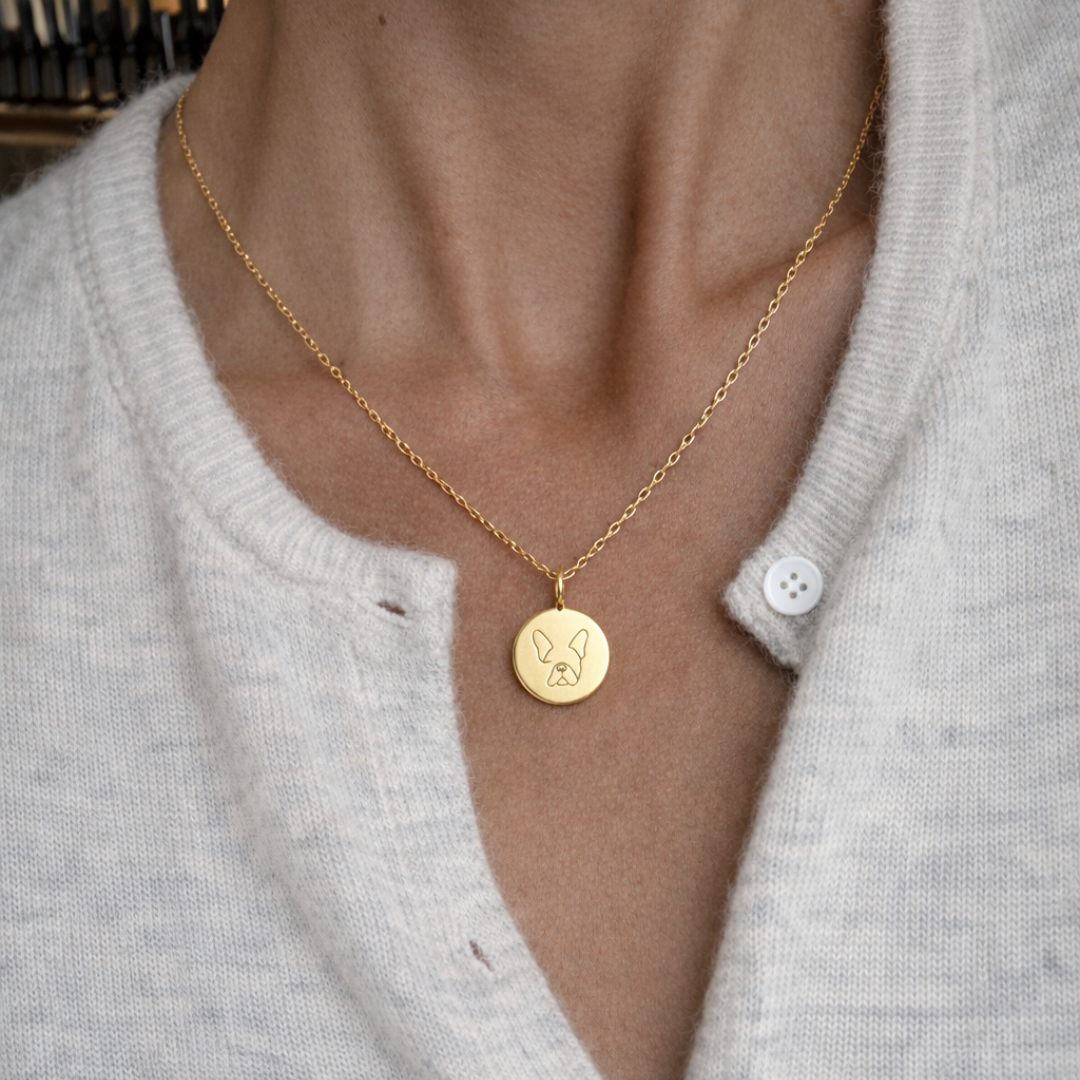 Custom engraved 18k gold vermeil 16mm single-line drawing coin pendant necklace worn at neckline on female model in cream cardigan, personalised minimalist French Bulldog face portrait with signature bat ears precision diamond-head engraved on warm lustrous gold disc on delicate diamond-cut cable chain – Atelier By Dsgn luxury custom pet portrait jewellery everyday wear gift for dog lovers, French Bulldog mum, pet memorial or new puppy celebration keepsake necklace