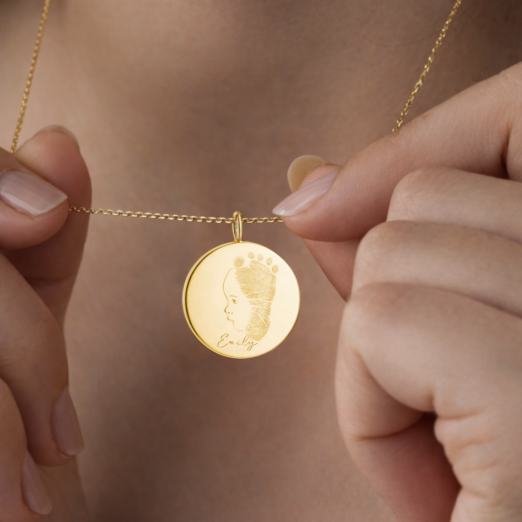 Female model holding custom engraved 18k gold vermeil 16mm coin pendant necklace at neckline, featuring personalised single-line minimalist baby side profile portrait fused with actual baby footprint texture and name "Emily" precision diamond-head engraved on warm lustrous gold disc on delicate gold cable chain – Atelier By Dsgn luxury custom fingerprint footprint baby portrait jewellery deeply personal gift for new mum, mothers day, baby shower or treasured newborn heirloom necklace