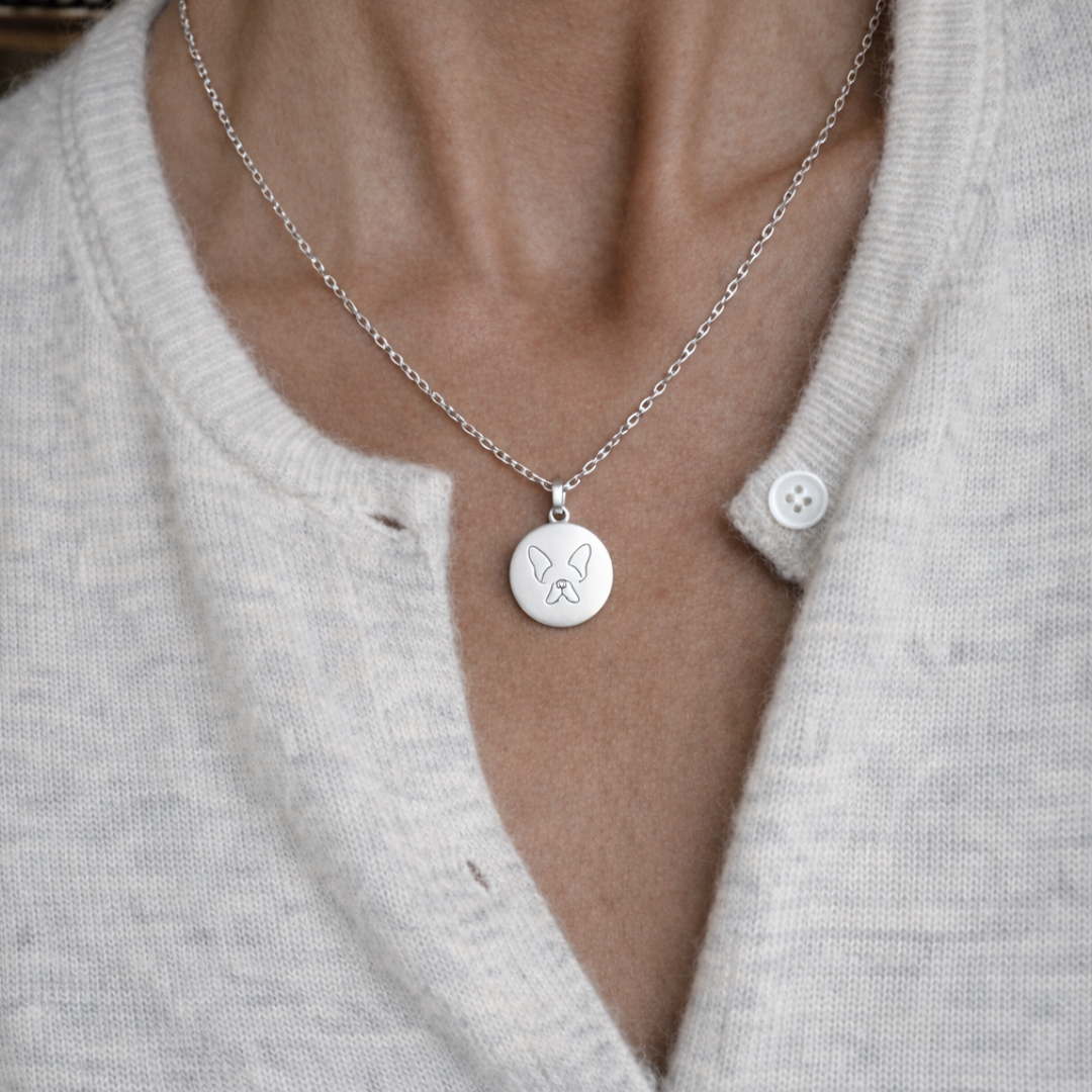 Custom engraved 925 sterling silver single-line drawing coin pendant necklace worn at neckline on female model in cream cardigan, personalised minimalist French Bulldog face portrait precision diamond-head engraved on polished round disc on delicate diamond-cut cable chain – Atelier By Dsgn custom pet portrait jewellery everyday wear gift for dog lovers, French Bulldog mum, pet memorial or new puppy celebration keepsake necklace