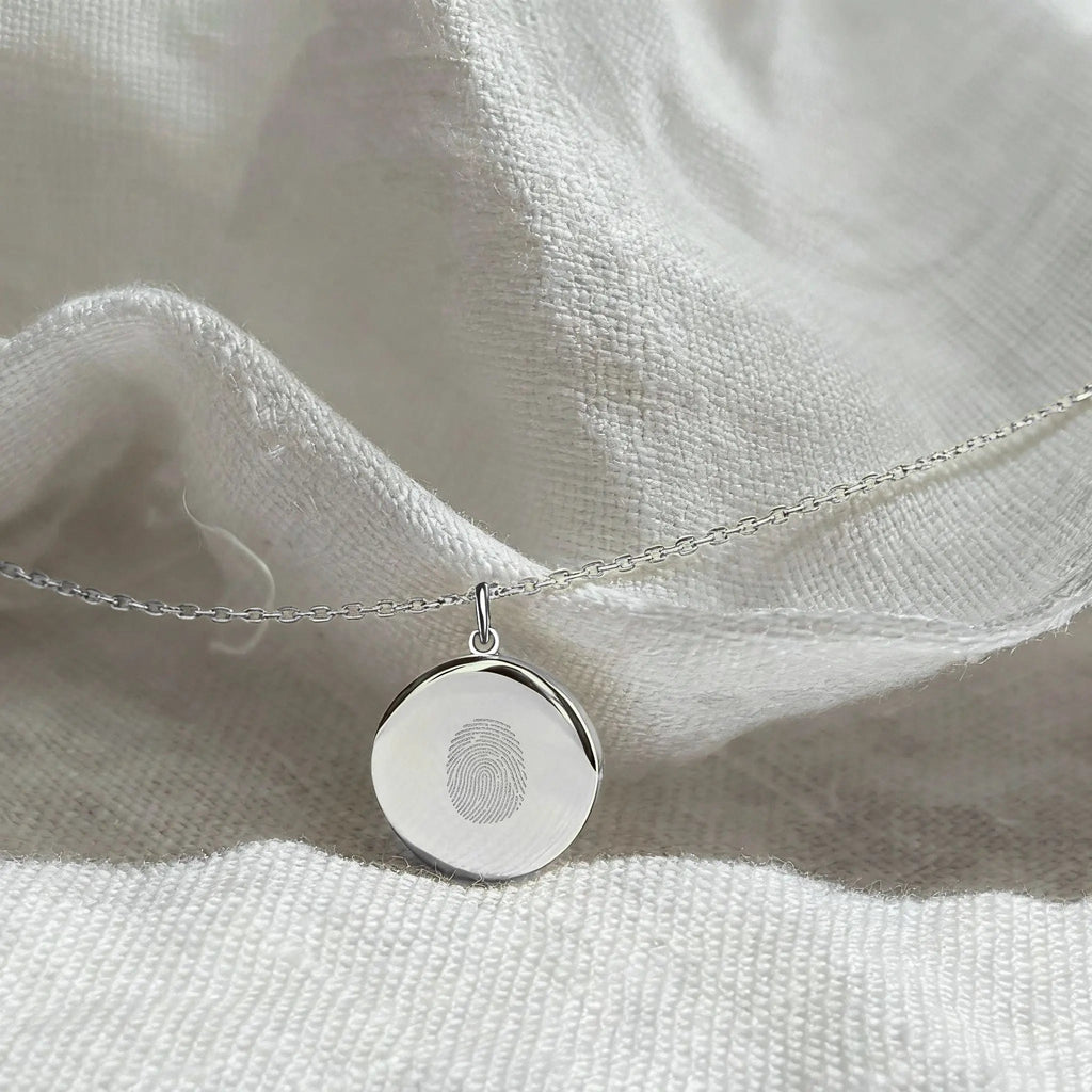 Close-up of the Atelier By DSGN Single Fingerprint Pendant Necklace in 925 Sterling Silver resting on soft white fabric showcasing the fine diamond-engraved fingerprint design and elegant minimalist craftsmanship of this personalized keepsake jewelry.
