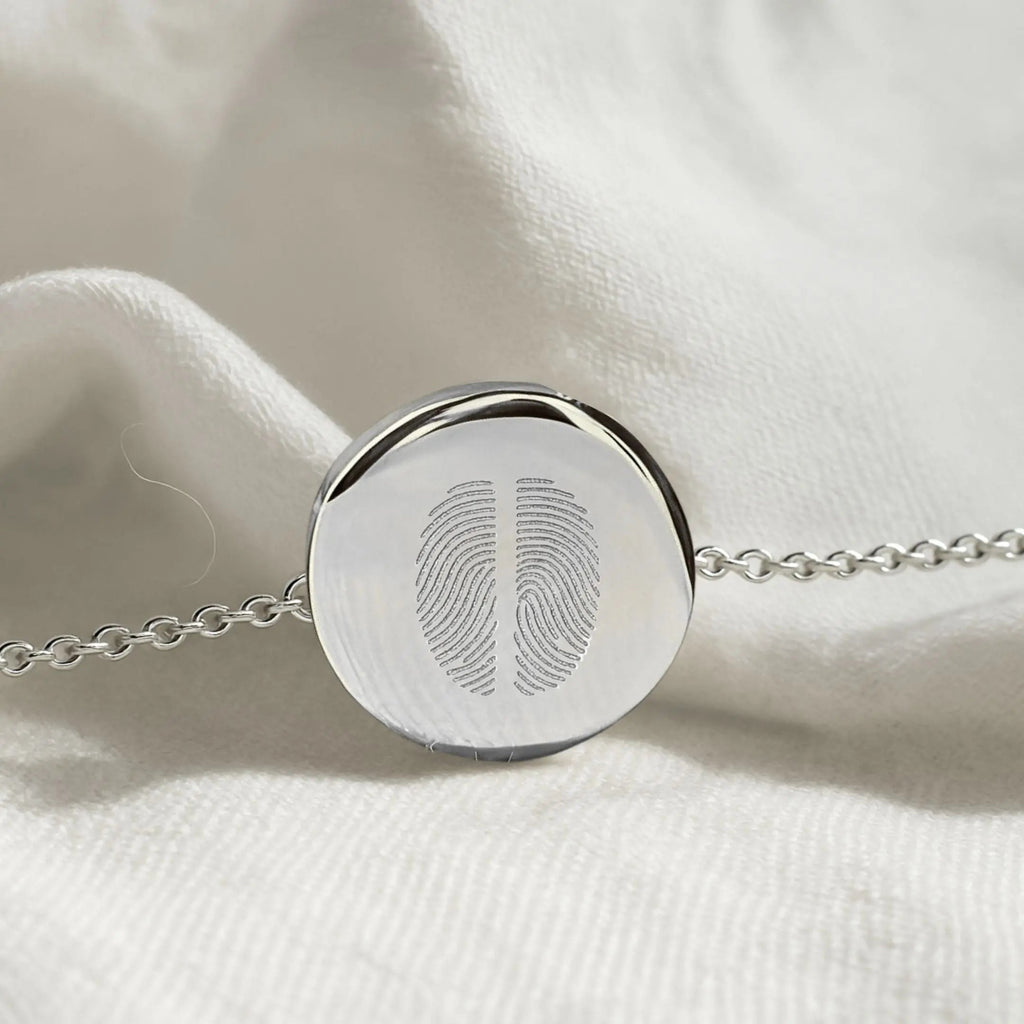 Close-up of the Atelier By DSGN Two Fingerprint Coin Bracelet in 925 Sterling Silver resting on soft white fabric, highlighting the dual fingerprint engraving and polished coin pendant detail.