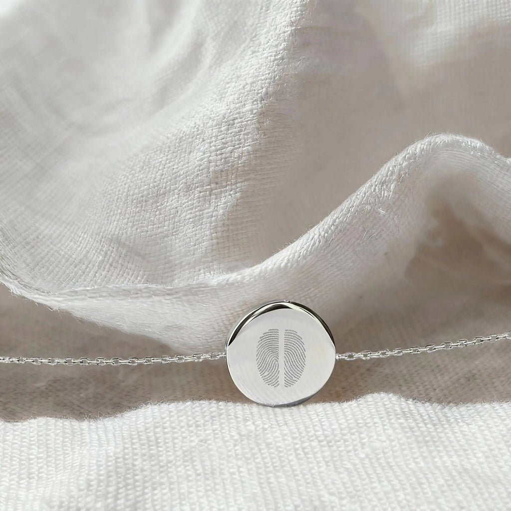 The Atelier By DSGN Two Fingerprint Coin Bracelet in 925 Sterling Silver resting on softly textured white fabric, highlighting the detailed dual fingerprint engraving and polished round pendant.