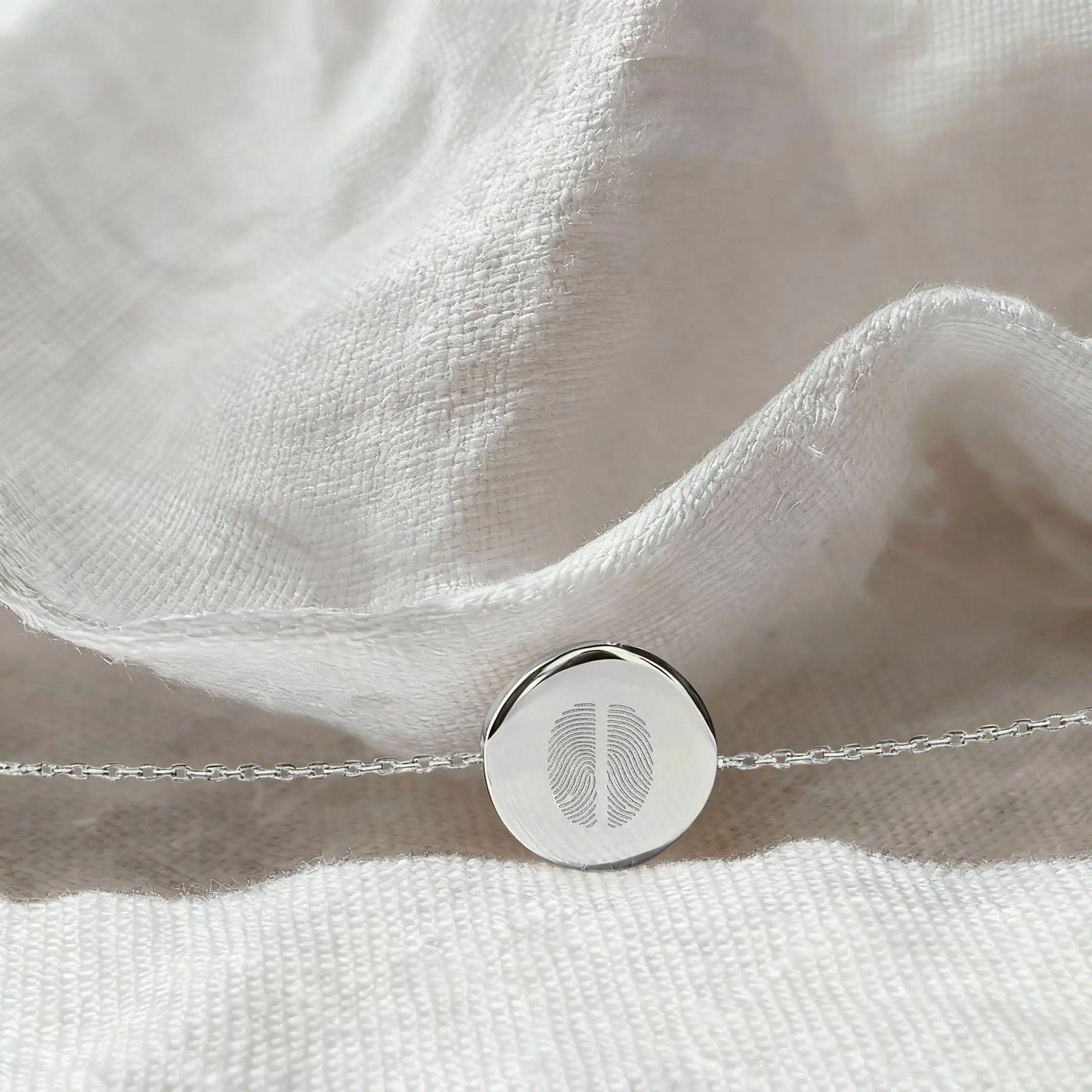 The Atelier By DSGN Two Fingerprint Coin Bracelet in 925 Sterling Silver resting on softly textured white fabric, highlighting the detailed dual fingerprint engraving and polished round pendant.