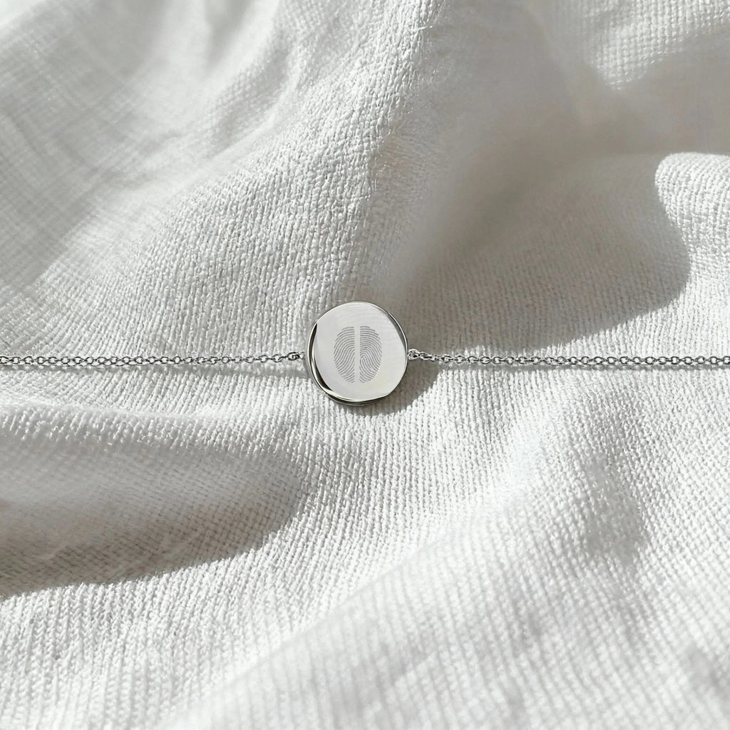 The Atelier By DSGN Two Fingerprint Coin Bracelet in 925 Sterling Silver displayed flat on soft white fabric, showcasing the delicate dual fingerprint engraving and elegant minimalist chain.