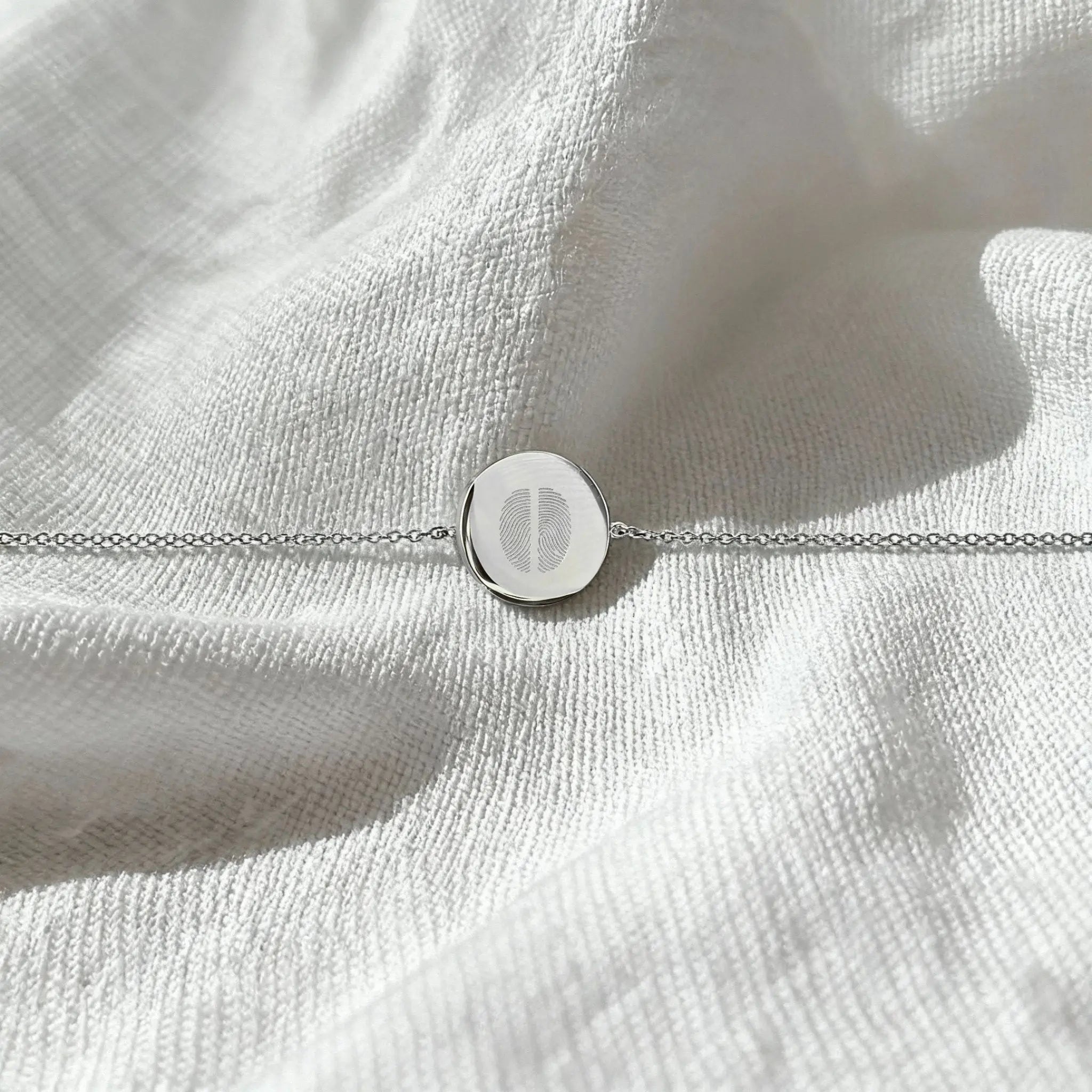The Atelier By DSGN Two Fingerprint Coin Bracelet in 925 Sterling Silver displayed flat on soft white fabric, showcasing the delicate dual fingerprint engraving and elegant minimalist chain.