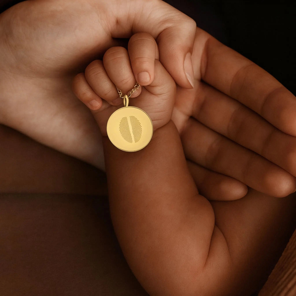 A tender close-up of a baby’s hand resting in their mother’s palm, both holding the  Atelier By DSGN Fingerprint Coin Necklace in 18K Gold Vermeil. The pendant features two fingerprints elegantly engraved together, symbolising the unbreakable bond between mother and child a timeless piece of love, memory, and connection.