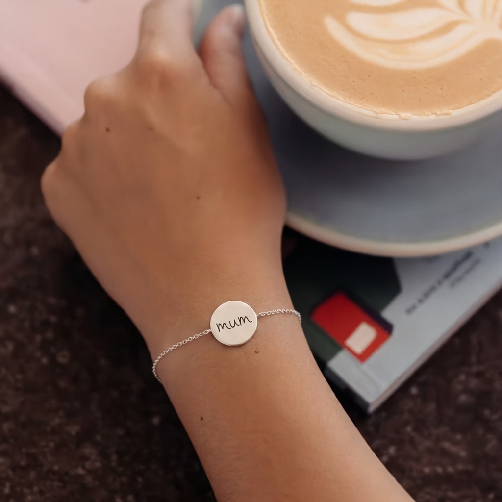 A sterling silver custom handwriting engraved chain bracelet with a round engraved disc that reads 'mum' worn on a person's wrist, with a hand holding a cup of latte in a cafe setting.  Custom meaningful jewelry from Atelier By DSGN.