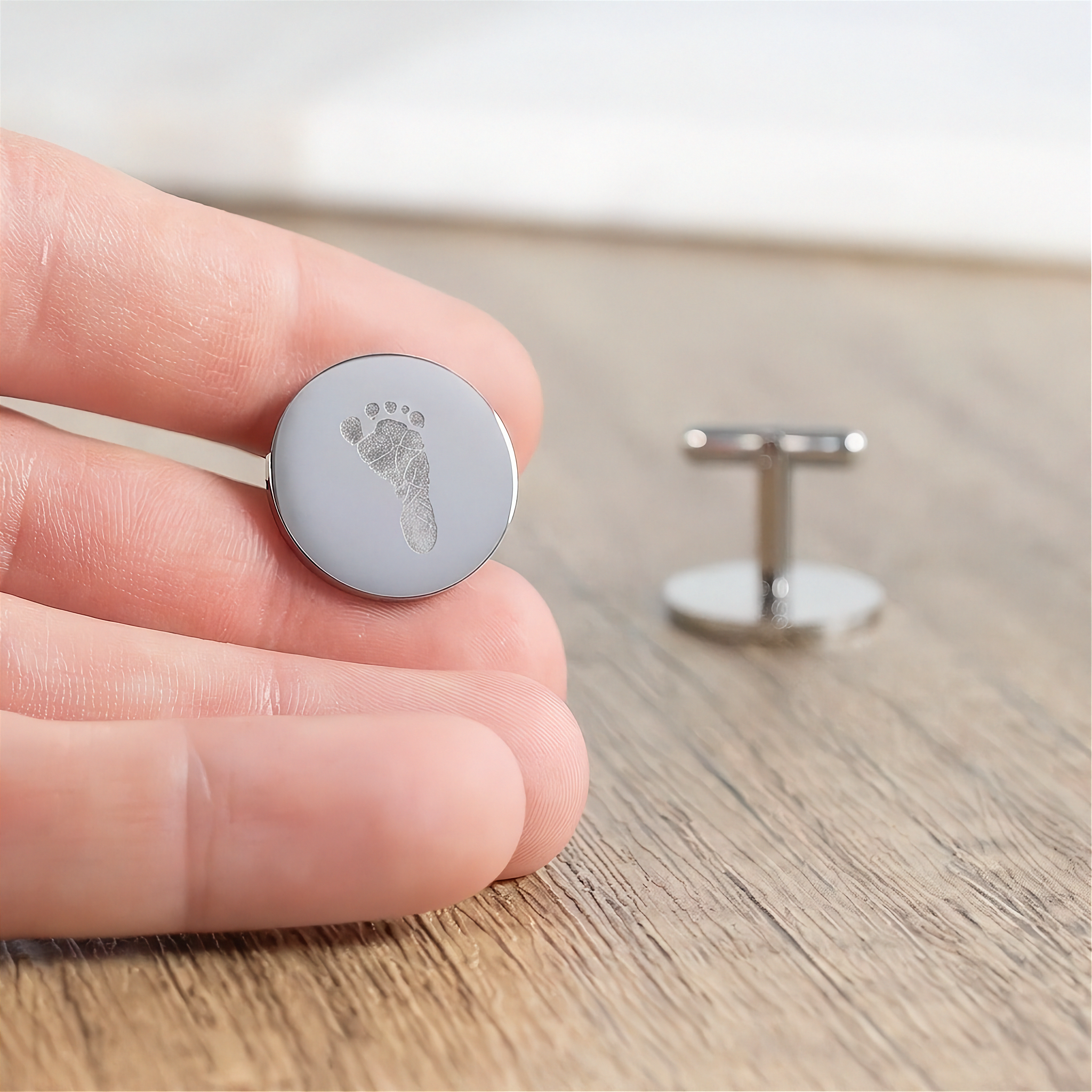 A close-up of a hand holding a round sterling silver cufflink engraved with a tiny baby footprint, with another cufflink blurred in the background on a wooden surface.  Personalised men's jewelry from Atelier By DSGN.