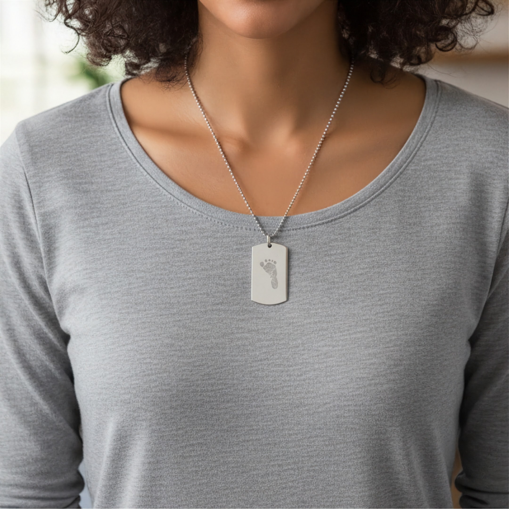 A close-up of a person wearing a silver tag pendant necklace custom engraved with a baby footprint, paired with a light gray long-sleeve top.  Personalised foot print engraved silver jewelry from Atelier By DSGN