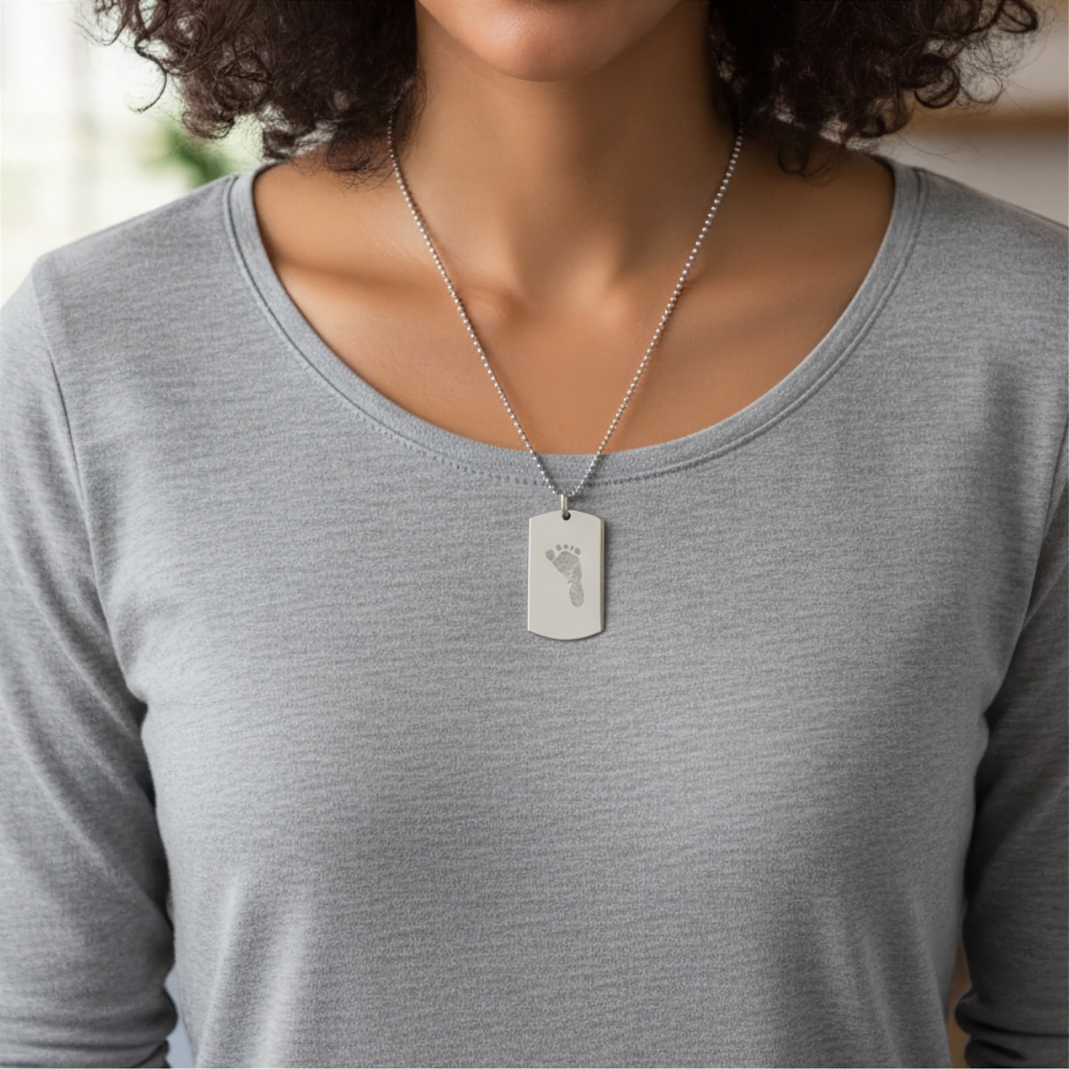 A close-up of a person wearing a silver tag pendant necklace custom engraved with a baby footprint, paired with a light gray long-sleeve top.  Personalised foot print engraved silver jewelry from Atelier By DSGN