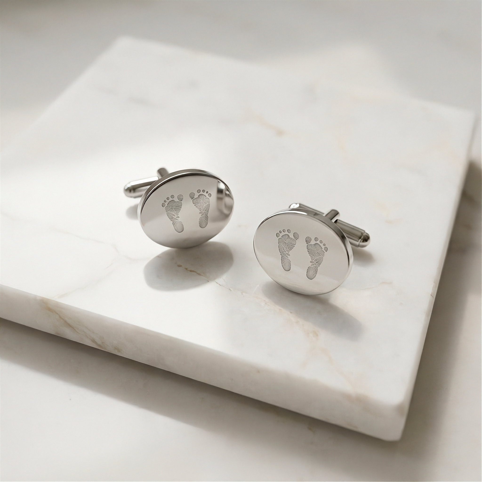 A pair of round coin sterling silver cufflinks engraved with two tiny footprints next to each other.  Cufflinks are resting on a white marble display table against a light gray background.  Custom jewelry from Atelier By DSGN.