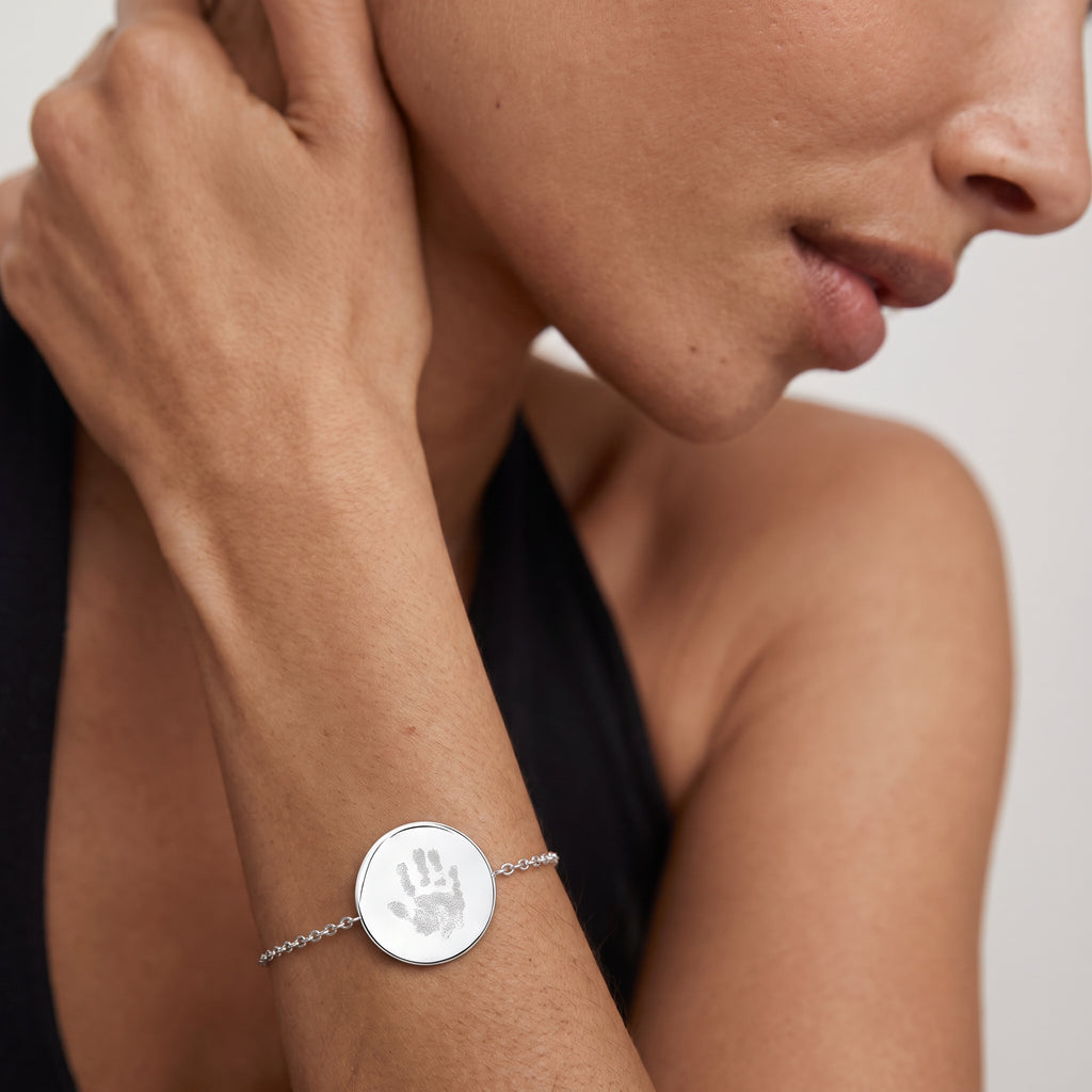 A close-up of a lady's wrist wearing a delicate silver chain , coin pendant bracelet with a round silver disc engraved with a childs' handprint.  Custom jewelry from Atelier By DSGN