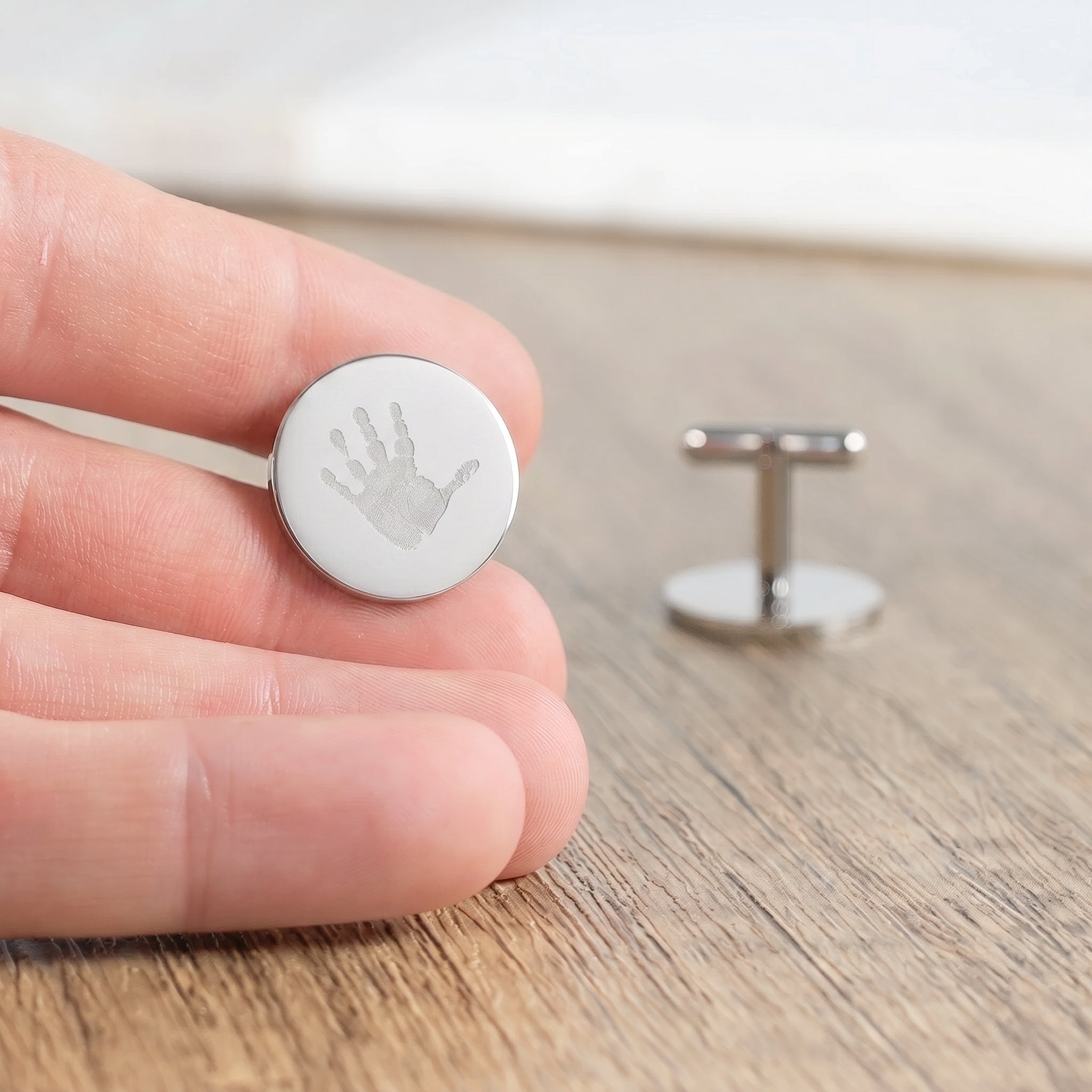 A close-up of a hand holding a round sterling silver cufflink engraved with a baby handprint, with the matching cufflink displayed on a wooden surface in the background.  Meaningful memorial jewelry from Atelier By DSGN.