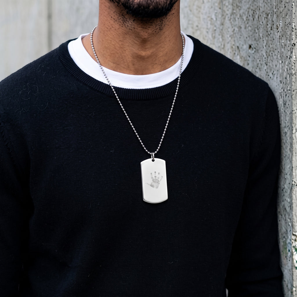 A 925 sterling silver ball chain necklace with a rectangular dog tag pendant featuring a custom engraved handprint, worn by a man in a black crewneck sweater over a white undershirt, standing against a textured gray concrete wall.  Meaningful jewelry from Atelier By DSGN.
