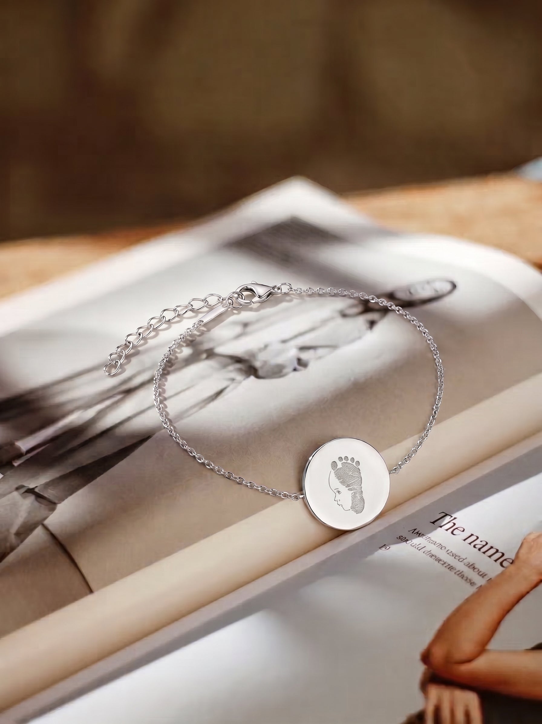 A close up of a delicate silver chain bracelet with a round engraved disc pendant, pendant is engraved with a single line portrait and print and the bracelet is displayed on an open magazine. Custom jewelry from Atelier By DSGN.