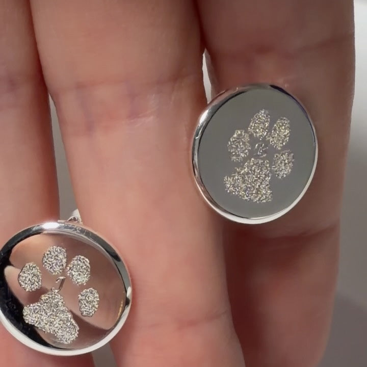 A close-up of a hand holding two round silver coin cufflinks with paw print designs engraved on them, one cufflink between each finger.