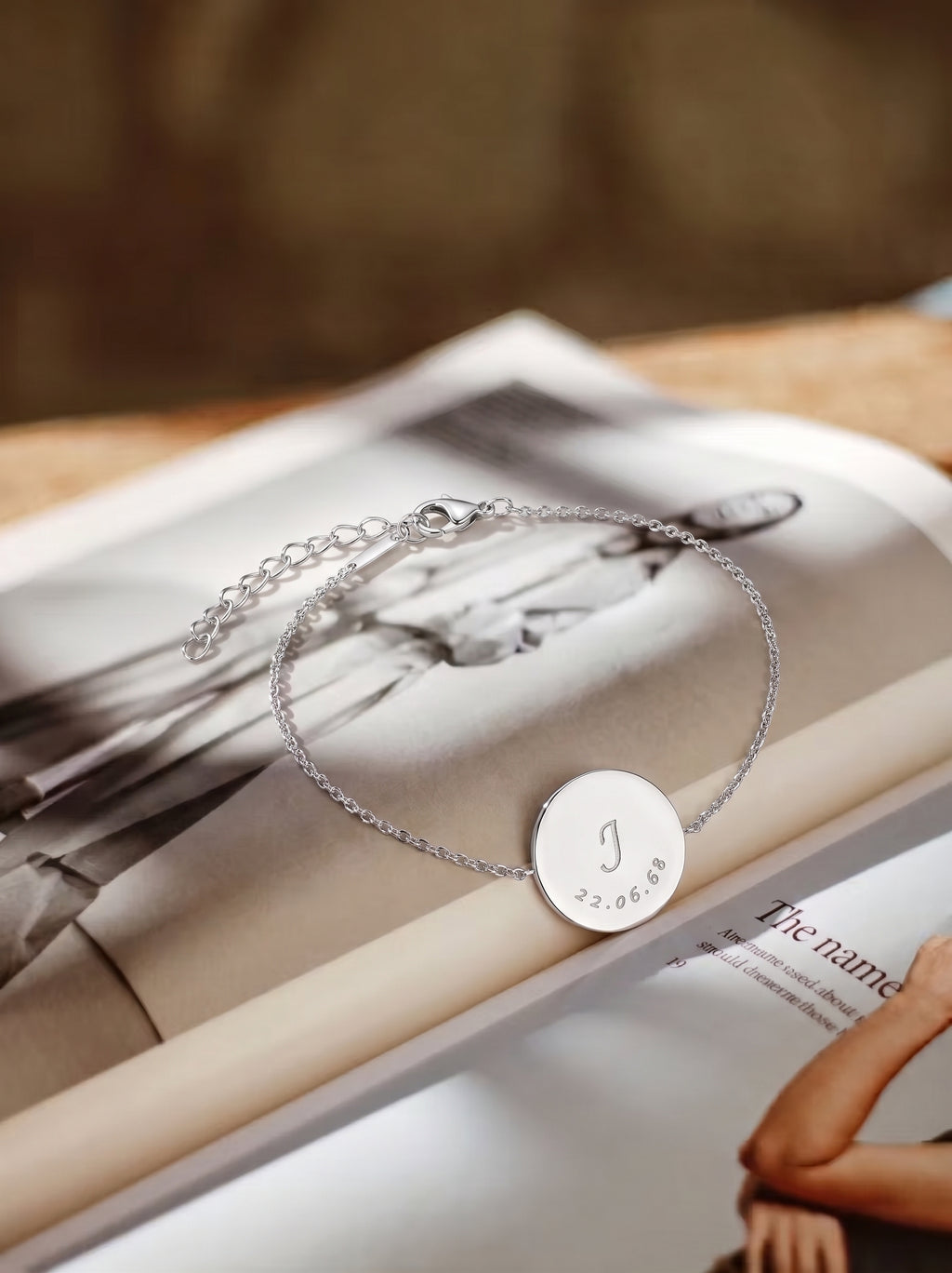 A delicate silver chain bracelet with a round engraved disc pendant, displayed on an open magazine. The pendant features a custom engraved monogram 'J' and the date '3.  Personalised jewelry from Atelier By DSGN.