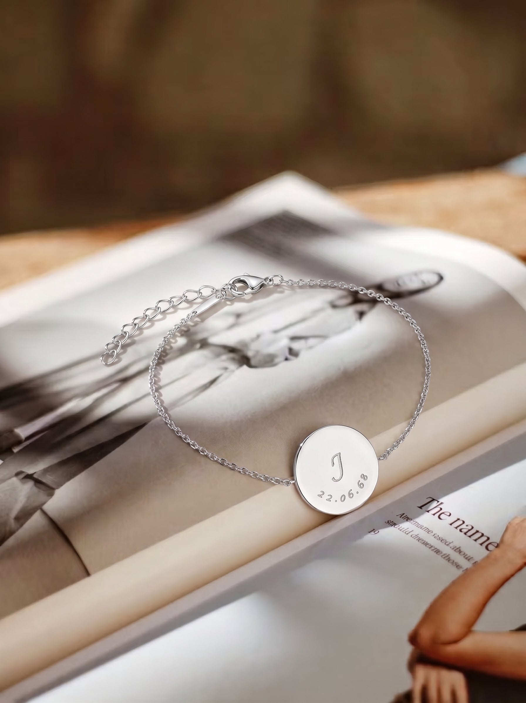 A delicate silver chain bracelet with a round engraved disc pendant, displayed on an open magazine. The pendant features a custom engraved monogram 'J' and the date '3.  Personalised jewelry from Atelier By DSGN.
