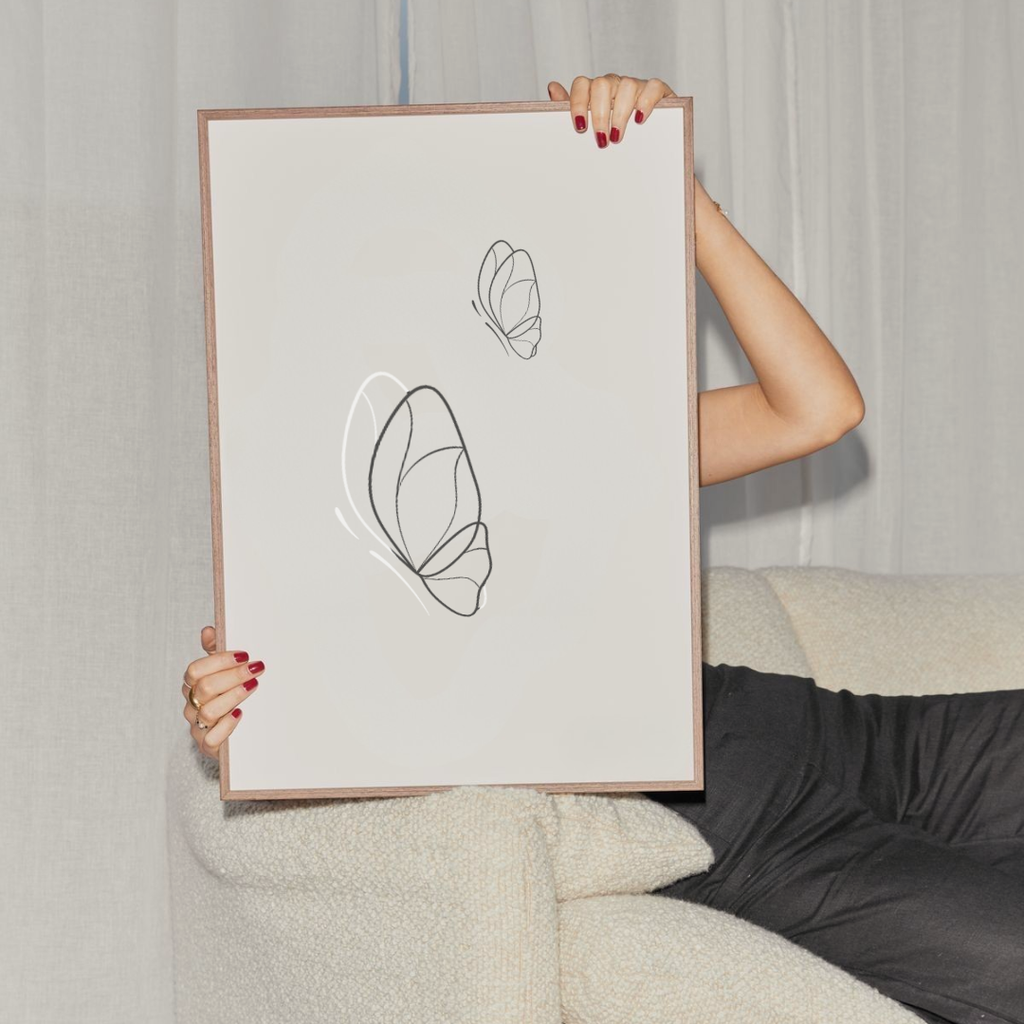 A person holds up a large framed minimalist art print featuring two butterfly line drawings  which is created from their pet's ear outline and turned into a butterfly design, on an off-white background. The print has a light wood frame and is shown in a cozy indoor setting with a textured cream sofa and neutral curtains in the background.