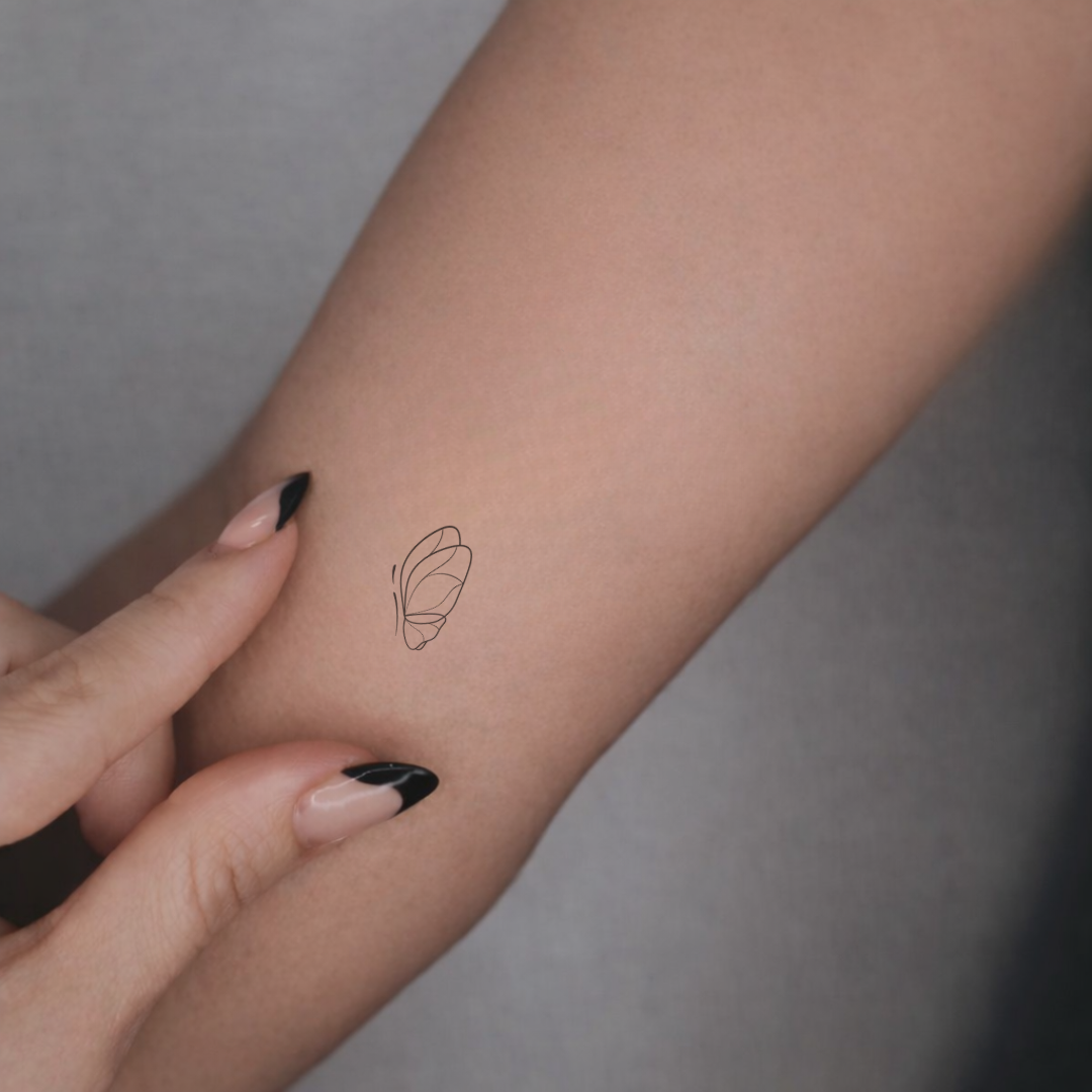 Three small minimalist pet feature butterfly design line-art used as a tattoos design on a person's forearm, shown with a hand pointing to them against a soft gray background.  Meaningful designs from Atelier By DSGN.