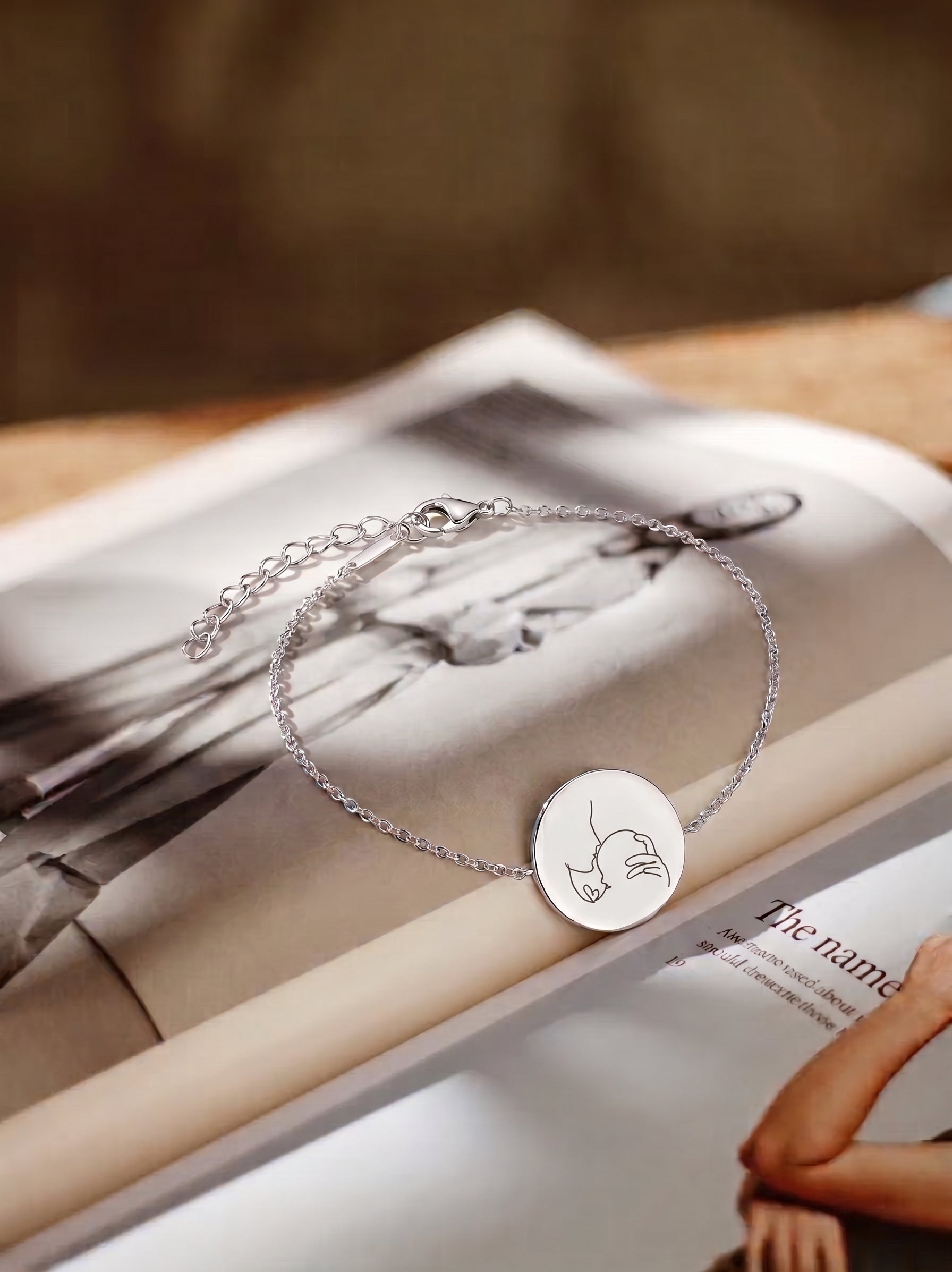 A close up of a delicate silver chain bracelet with a round engraved disc pendant, displayed on an open magazine. The pendant features a custom engraved single line portrait of a mother and her baby with a heart in the middle.  Custom jewelry from Atelier By DSGN. 