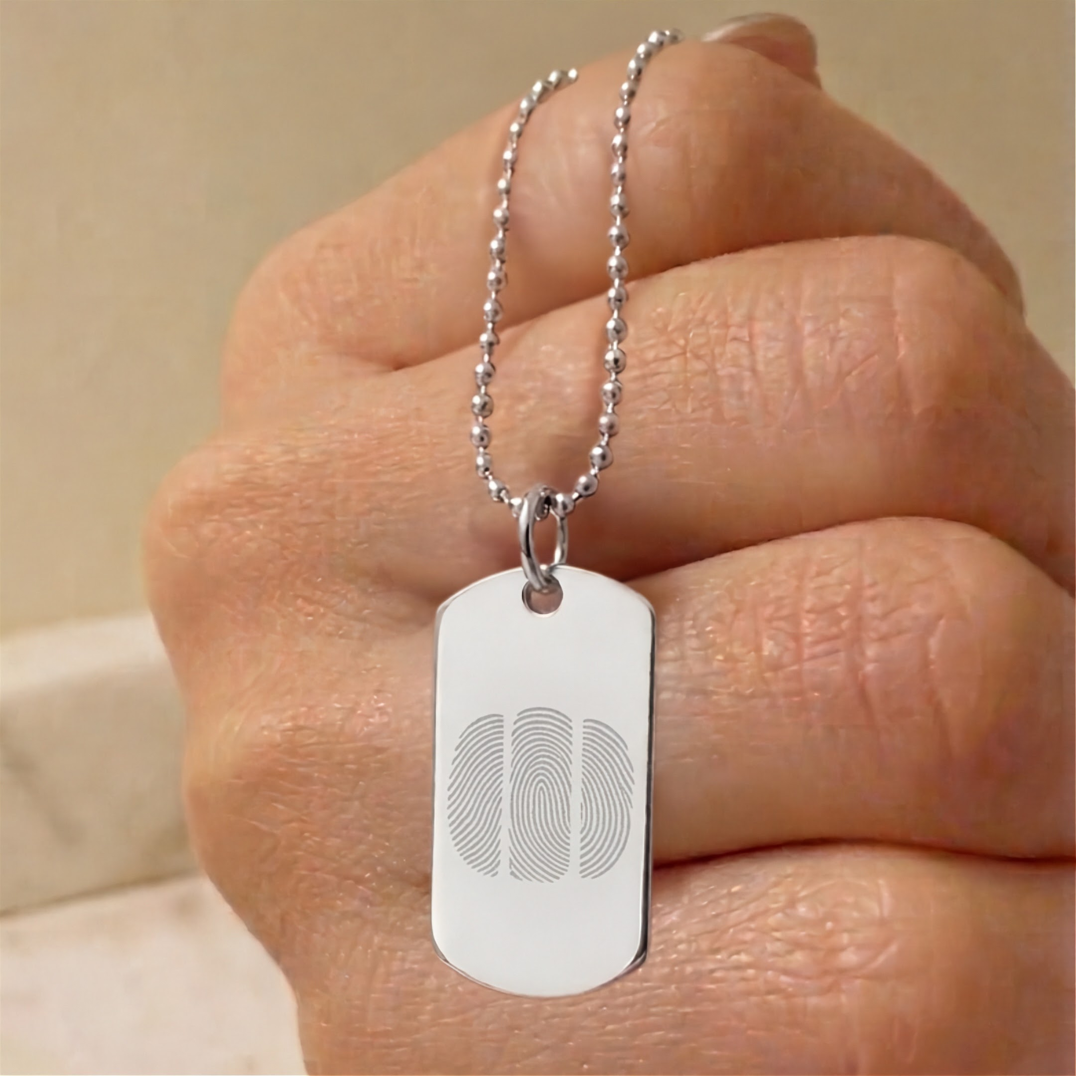 A silver dog tag necklace with a ball chain, held in a hand. The dog tag features an engraved three fingerprint design.  Custom jewelry from Atelier By DSGN