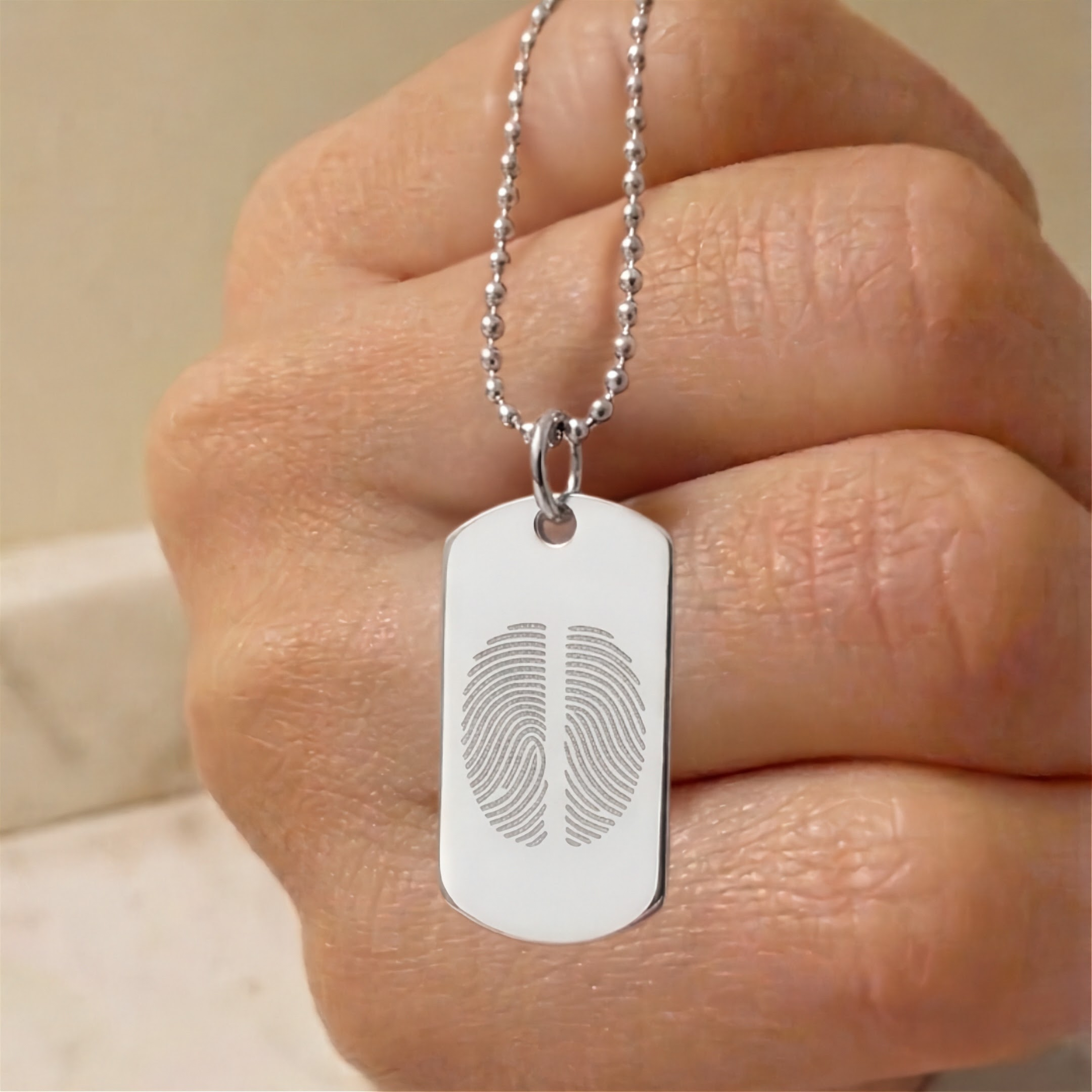 A close-up of a hand holding a silver ball-chain necklace with a dog tag pendant engraved with two fingerprint designs. Custom jewelry from Atelier By DSGN.