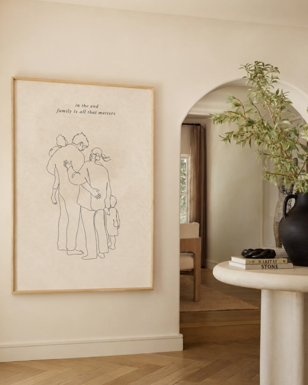 Large, framed single line family drawing print from Atelierbydsgn displayed in a modern beige living room. Features a minimalist personalized family portrait illustration with text, styled for minimalist home decor in a wooden frame.