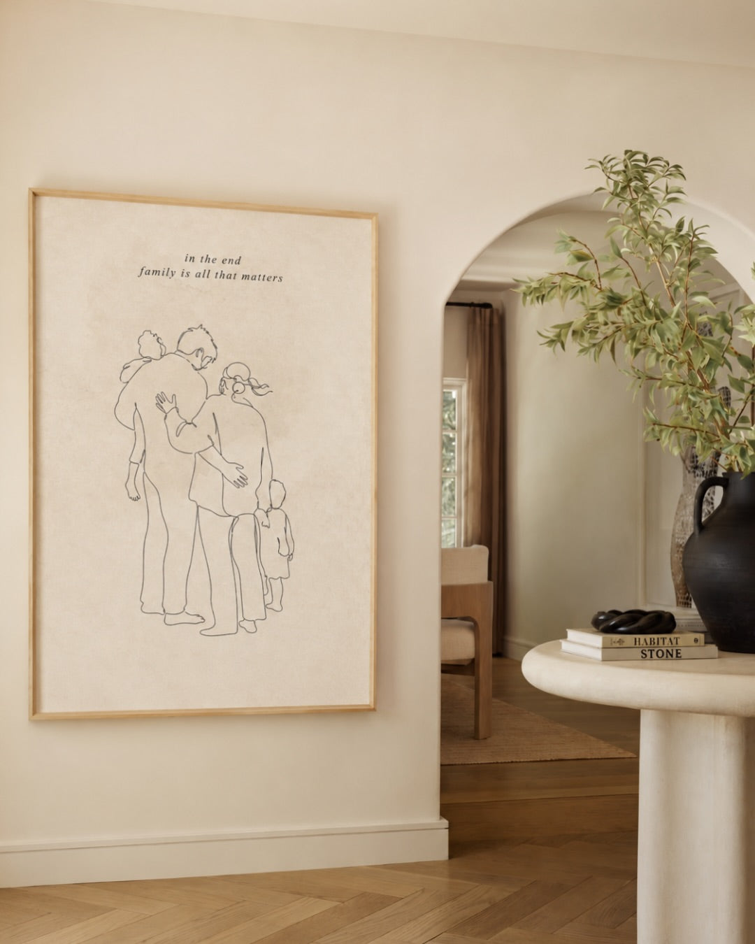 Large, framed single line family drawing print from Atelierbydsgn displayed in a modern beige living room. Features a minimalist personalized family portrait illustration with text, styled for minimalist home decor in a wooden frame.