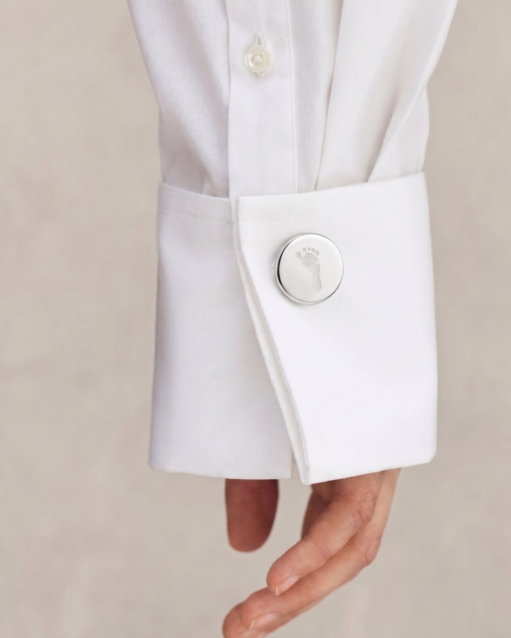 Close-up of a white dress shirt sleeve with a single decorative round footprint engraved cufflinks on the cuff, worn on a person's arm against a soft beige background.  Custom jewelry from Atelier By DSGN
