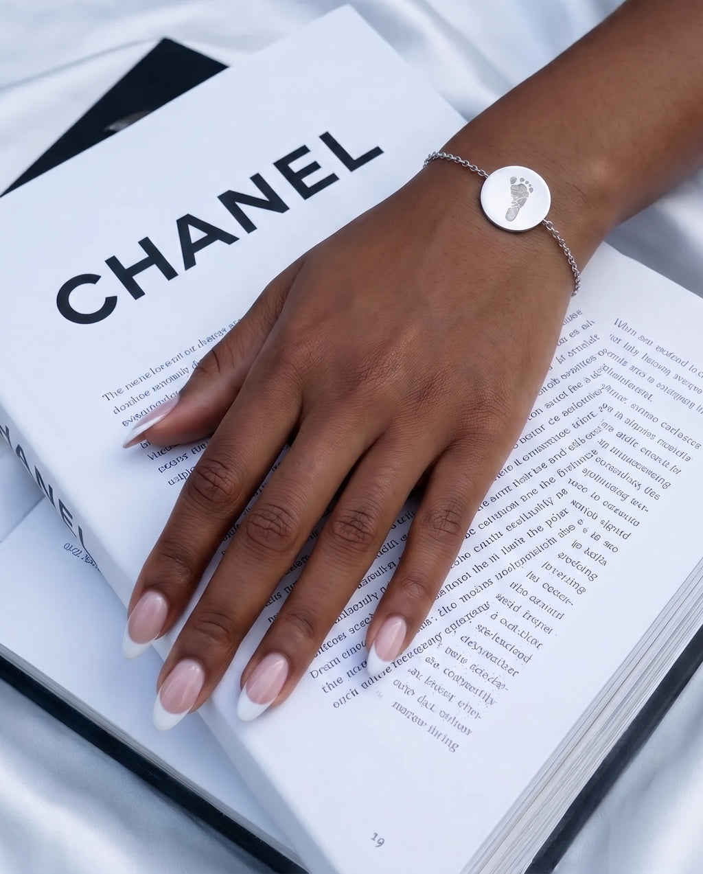 Atelier By DSGN custom footprint engraved 925 sterling silver bracelet on a lady's wrist.  Lady has french manicured nails and her hand is resting on a Chanel book.  Delicate meaningful jewelry. 