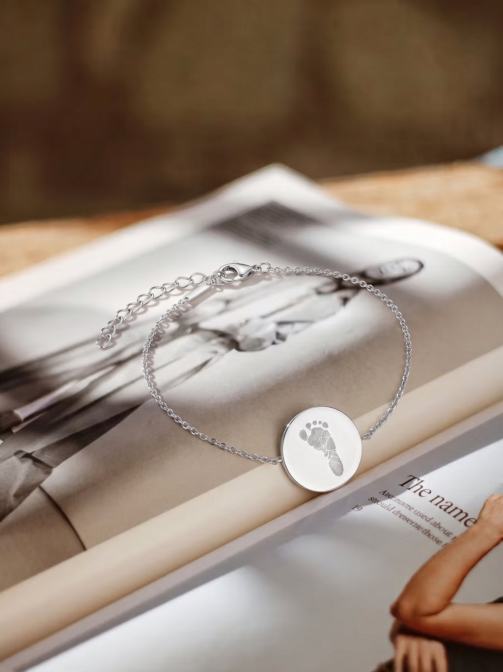 Custom footprint engraved sterling silver bracelet laying on a book.  Shows the close up of the delicate foot print engraving on the coin pendant.  Custom jewelry from Atelier By DSGN.