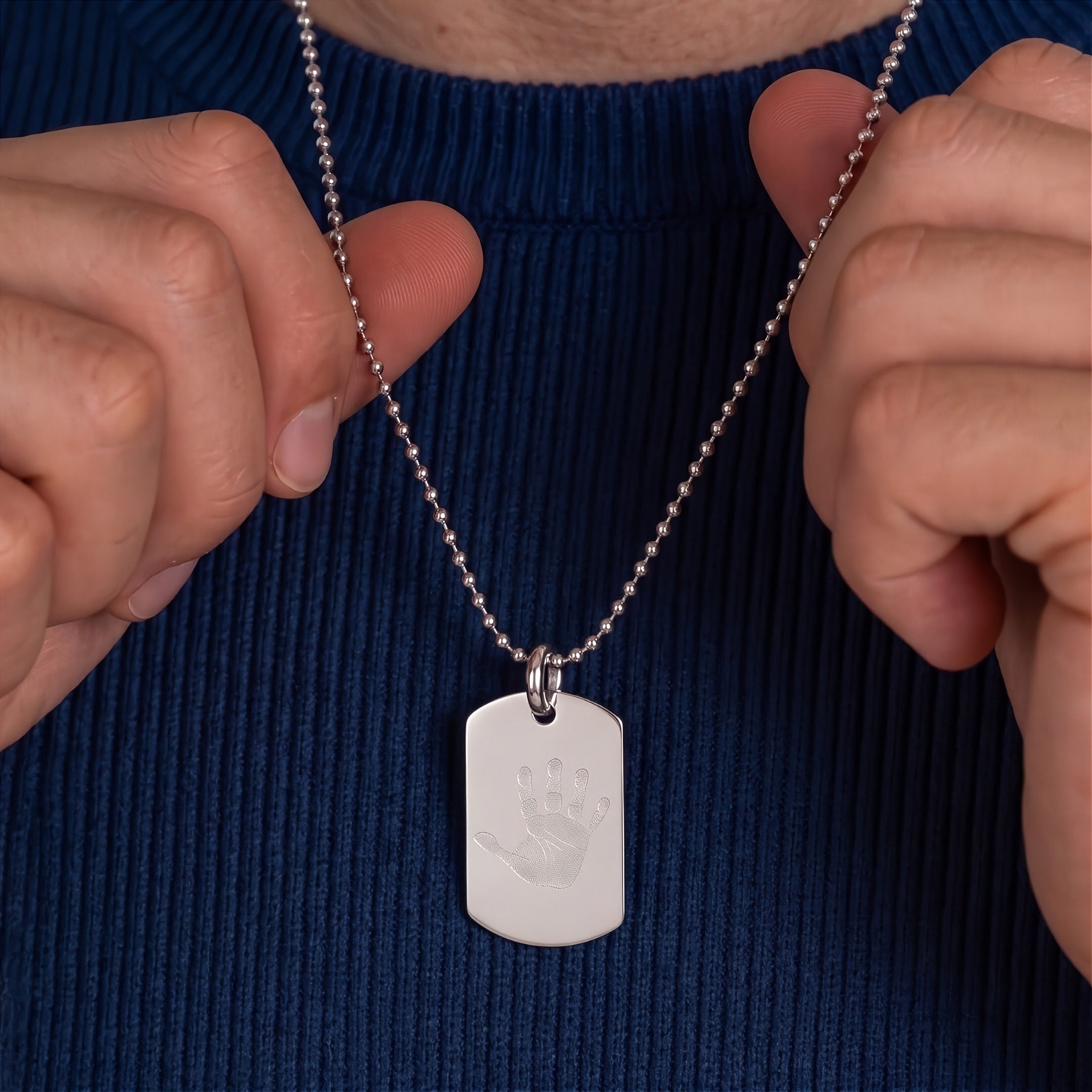 A 925 sterling silver dog tag necklace with an engraved handprint print, around the neck of a man held by a person wearing a dark blue ribbed sweater.  Luxury jewelry from atelier by dsgn,