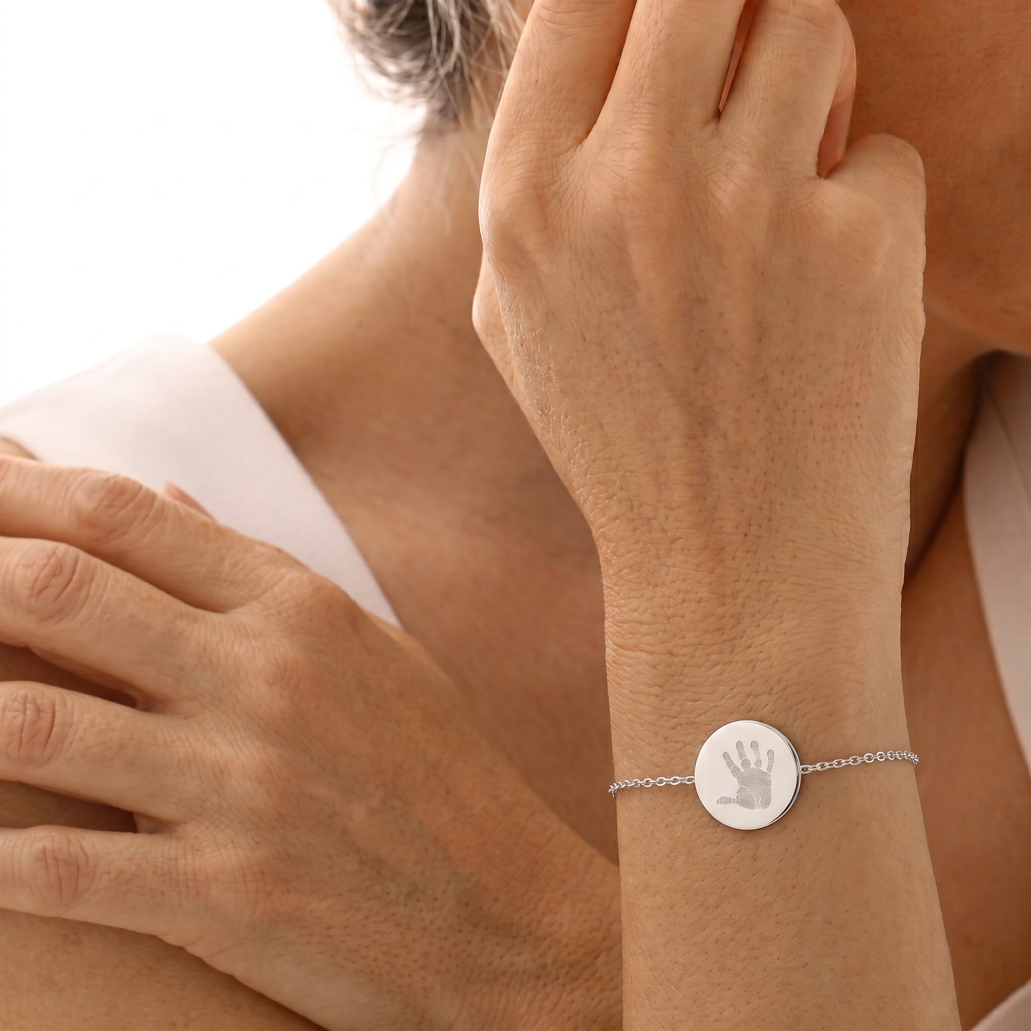 A close-up of a person's wrist wearing a delicate silver chain bracelet with a round silver disc engraved with a handprint.  Custom jewelry from Atelier By DSGN