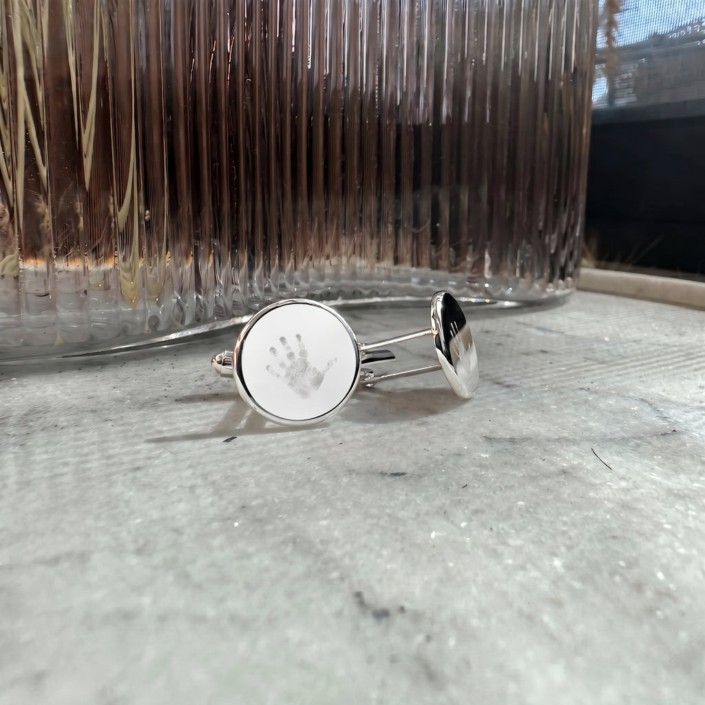 A close up of a pair of silver circle cufflinks engraved with a handprint and resting on a gray marble surface in front of a ribbed glass vase. Custom jewelry from Atelier By DSGN