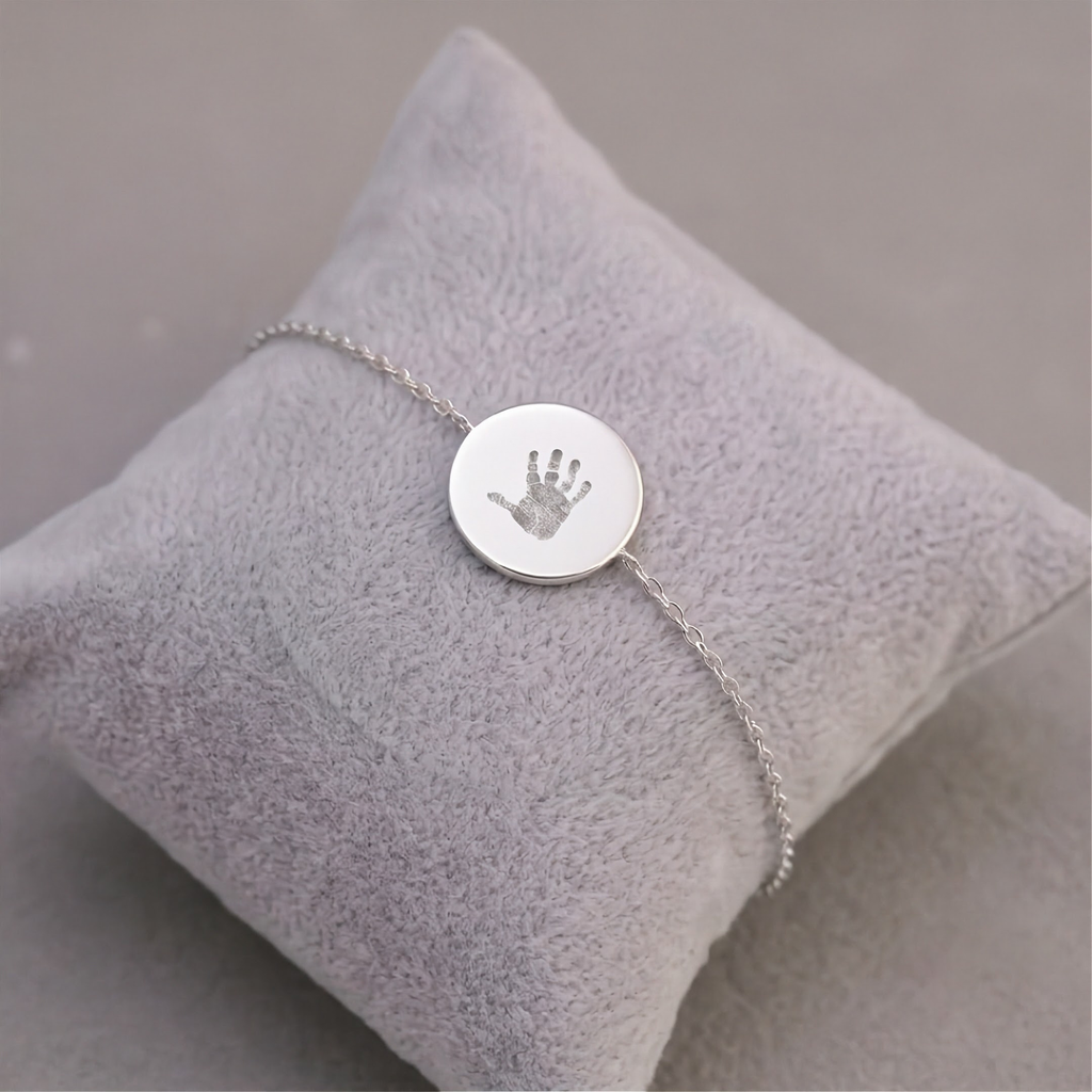 A delicate silver bracelet with a round disc charm engraved with a baby handprint, displayed on a soft, light gray velvet jewelry pillow.  Meaningful custom jewelry from Atelier By DSGN