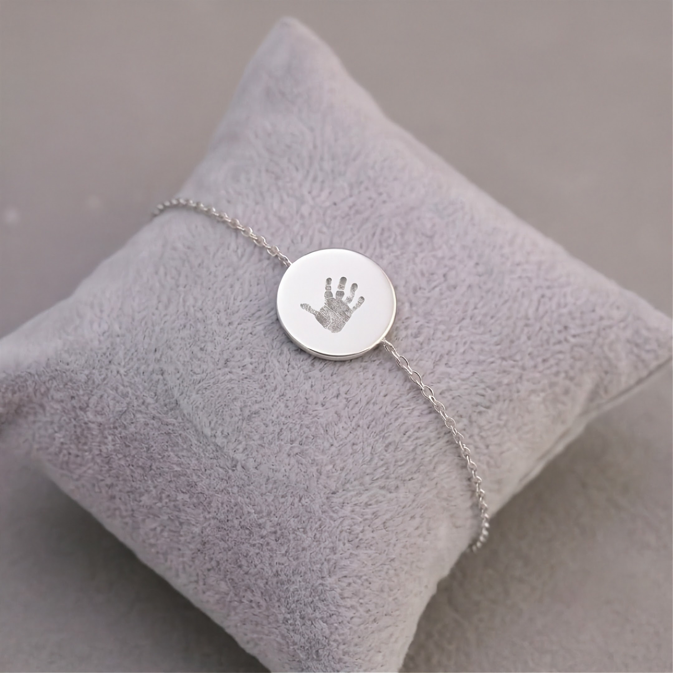 A delicate silver bracelet with a round disc charm engraved with a baby handprint, displayed on a soft, light gray velvet jewelry pillow.  Meaningful custom jewelry from Atelier By DSGN
