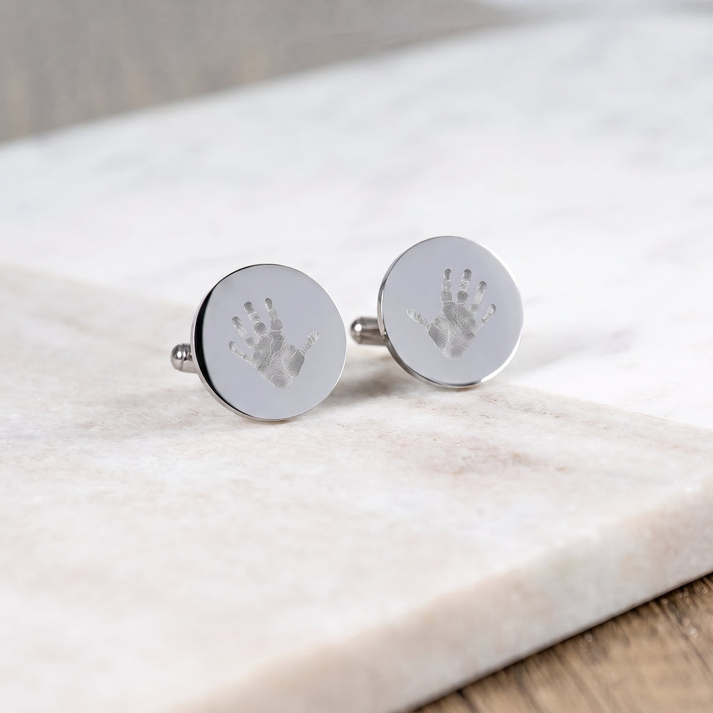 A pair of sterling silver coin cufflinks with small handprints engraved on each cufflink.  Cufflinks are sitting on a white marble table.  Custom men's jewelry from Atelier By DSGN.
