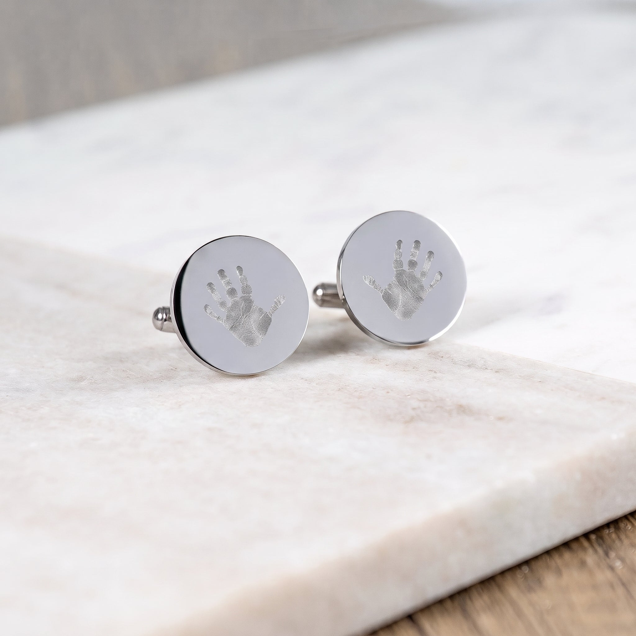A pair of sterling silver coin cufflinks with small handprints engraved on each cufflink.  Cufflinks are sitting on a white marble table.  Custom men's jewelry from Atelier By DSGN.
