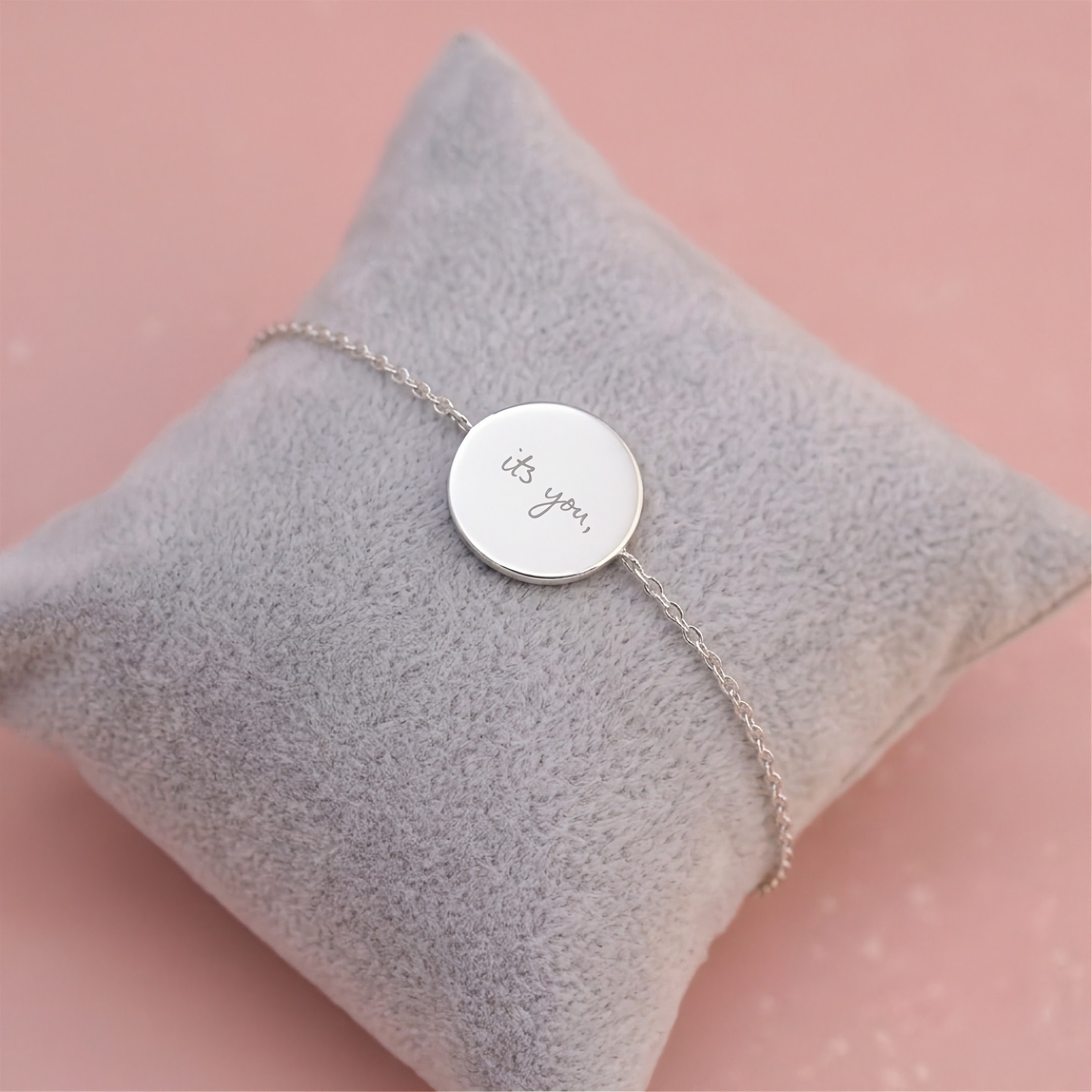 A delicate custom handwriting engraved silver coin bracelet and chain engraved with handwritten 'it's you,' displayed on a soft gray velvet jewelry pillow against a pale pink background.