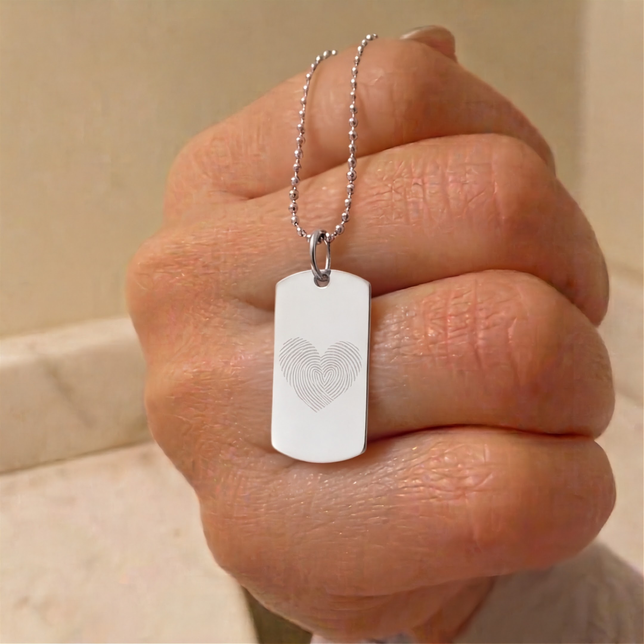 A close-up of a hand holding a silver ball chain necklace with a rectangular dog tag pendant engraved with concentric fingerprint heart shape.  Custom meaningful jewelry from Atelier By DSGN.