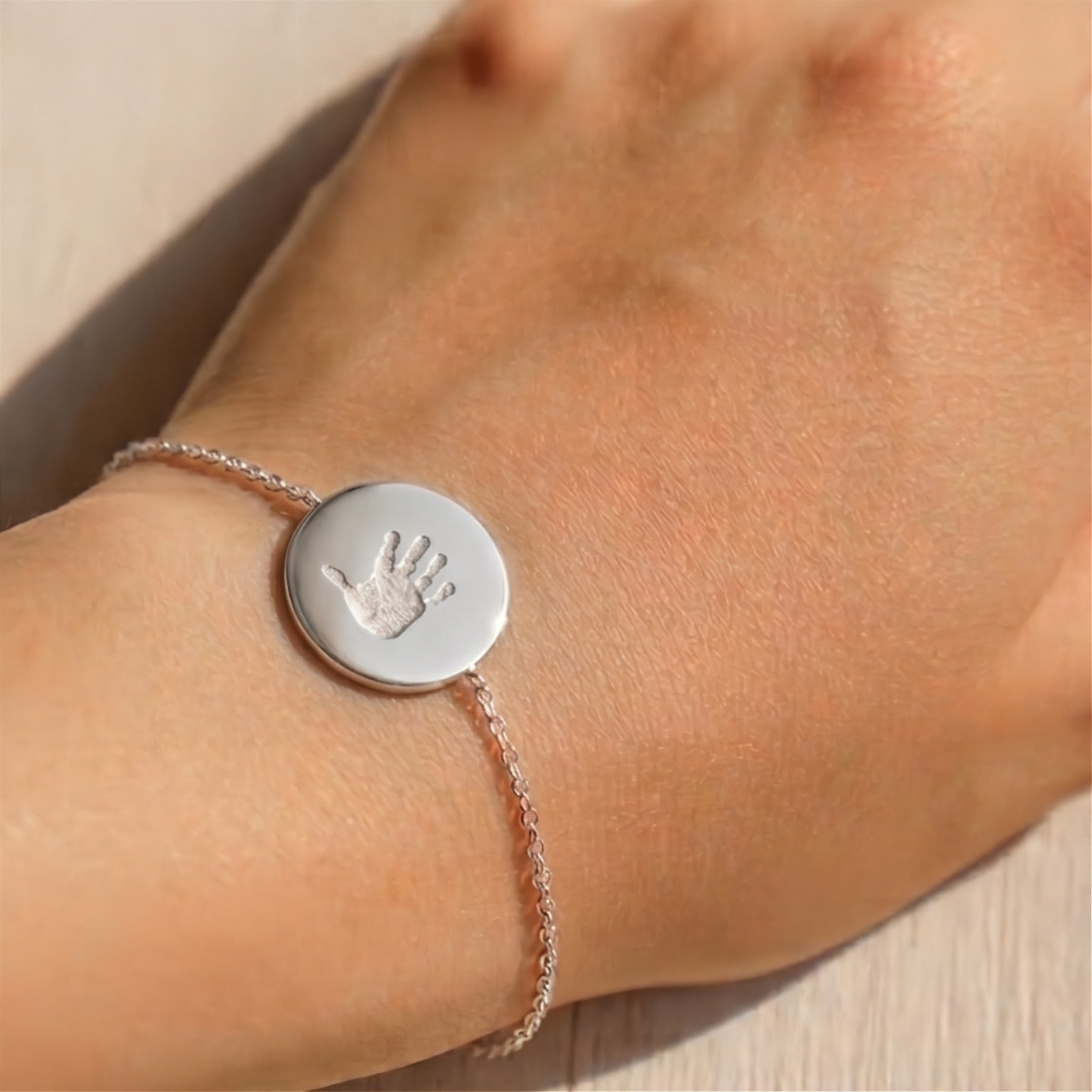 A close-up of a delicate silver bracelet worn on a wrist, featuring a round disc engraved with a baby handprint design.  Custom personal jewelry from Atelier By DSGN.