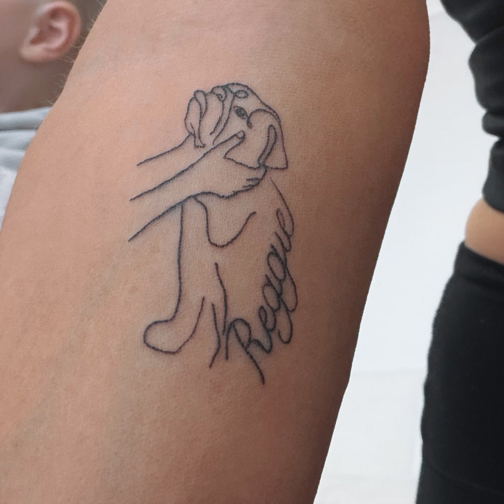 A custom line portrait tattoo of a bull dog and the owner's hands holding the dogs face tattooed on the inner arm with the ord Reggie on the side of the image.  Image designed by Atelier By DSGN.